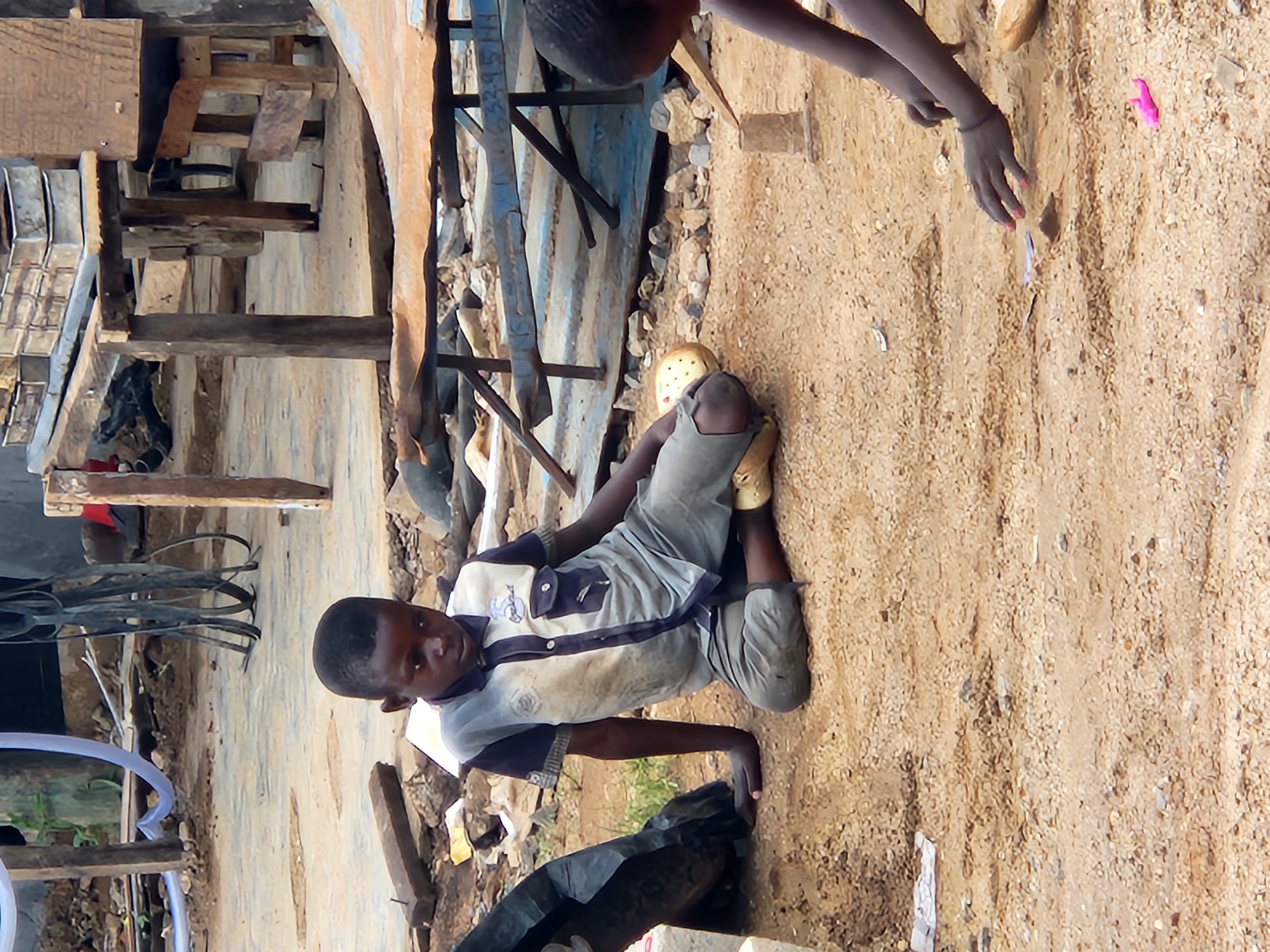 In Kabong, Nigeria, two young boys are observed in a dusty outdoor setting. The boy on the left, wearing a gray and navy collared shirt and gray pants, sits with his legs extended. He appears to be resting his right arm on his left leg, and his left hand is extended towards the ground. He is wearing yellow sandals. The second boy, on the right, is partially visible and is kneeling, reaching towards the ground with his hands. In front of the kneeling boy, there is a small pink object, possibly a toy, and a small structure made of mud or dirt. The environment is characterized by rough, sandy terrain with scattered debris, including pieces of wood, metal, and plastic. In the background, there are rudimentary wooden structures, suggesting a makeshift market or dwelling area. The overall scene depicts children engaged in play or an activity in a modest, possibly impoverished, urban environment.