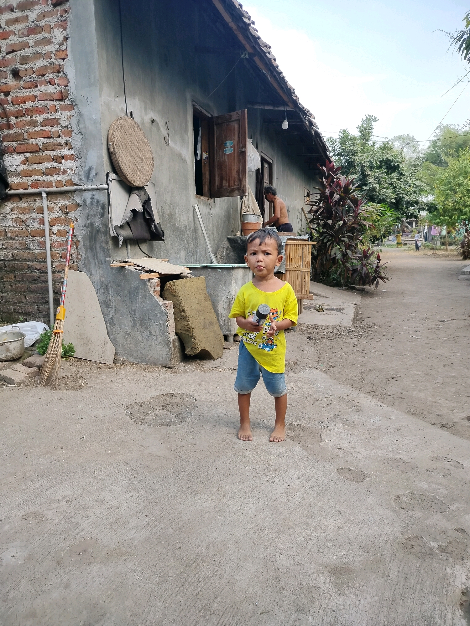 The image depicts an outdoor scene in the city of Beloh, Indonesia, featuring a young boy standing on a concrete path in front of a building. The boy, who appears to be around 5-7 years old, is wearing a yellow t-shirt with cartoon graphics and blue denim shorts. He is holding a black cylindrical object in his right hand and is looking directly at the camera. His bare feet are visible on the ground.
The building behind him is constructed with a combination of rough brickwork on the left and grey plastered walls on the right. An open wooden doorway is visible, with a person, seemingly an adult male, partially visible inside, shirtless and engaged in an activity. A woven circular object, possibly a basket or mat, hangs on the wall to the left of the doorway. A broom with a yellow handle is leaning against the wall on the far left of the image.
The environment appears to be a rural or semi-urban area, with a dirt path extending into the background, flanked by vegetation and other structures. The sky is visible overhead, with scattered clouds. The overall impression is one of a casual, everyday setting in a community.