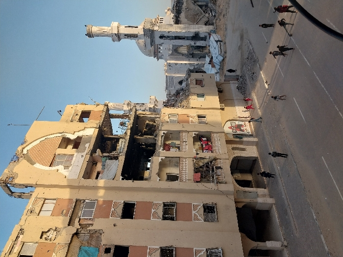 This image depicts a scene of extensive structural damage in Khan Yunis, Palestinian Territory. In the foreground and dominating the left side is a multi-story building, approximately six to seven floors high, exhibiting severe destruction. Its facade is heavily damaged with large sections missing, exposing interior rooms and structural elements. The upper floors are particularly compromised, with an ornate arched window feature on the top floor partially collapsed. Debris, exposed rebar, and charred interiors are visible on multiple levels, suggesting fire damage in some sections. Laundry is hanging on a balcony railing on the third floor, indicating recent or ongoing human presence. The ground floor features arched entrances and a visible sign with red lettering.
To the right of this building, a large mosque with a prominent minaret and dome is visible, also showing significant damage. Parts of its structure appear to be collapsed or heavily fragmented, and a large pile of rubble is present between the two main structures.
On the paved road and sidewalk in the lower half of the image, approximately ten to twelve individuals are scattered. Several children, some dressed in red, are visible closer to the left building, while a group of adults or older youths are further to the right on the street, some appearing to be walking or observing. The street itself has visible lane markings. The sky is clear and blue, and the lighting suggests late afternoon or early morning sun, casting gentle shadows. The overall environment conveys widespread devastation and disruption.