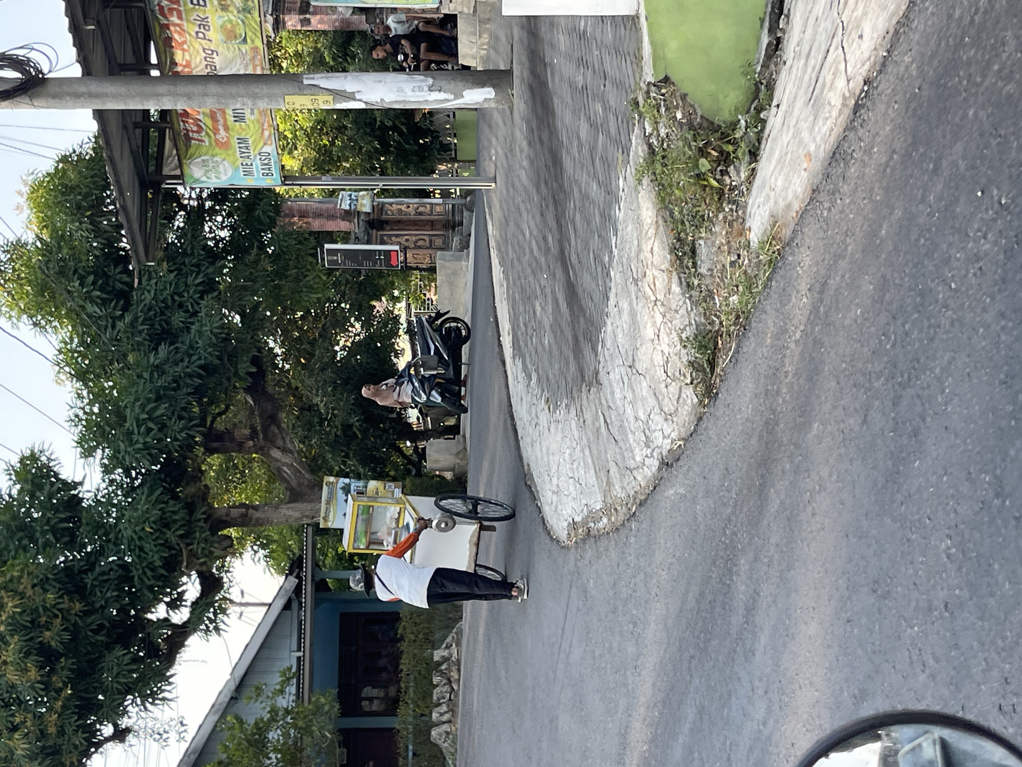 A person is pushing a food cart on a paved road in front of a building. The food cart has a yellow canopy with a clear front display case. The person is wearing a white shirt, orange sleeves, black pants, and a hat. A woman is sitting on a motorcycle to the right of the food cart, and another person is sitting on a step in the background. A large tree is visible behind the building. Signs are posted on a pole near the building, one of which displays "MIE AYAM" and "BAKSO". The environment appears to be a street scene in the city of Wonosegoro, Indonesia.