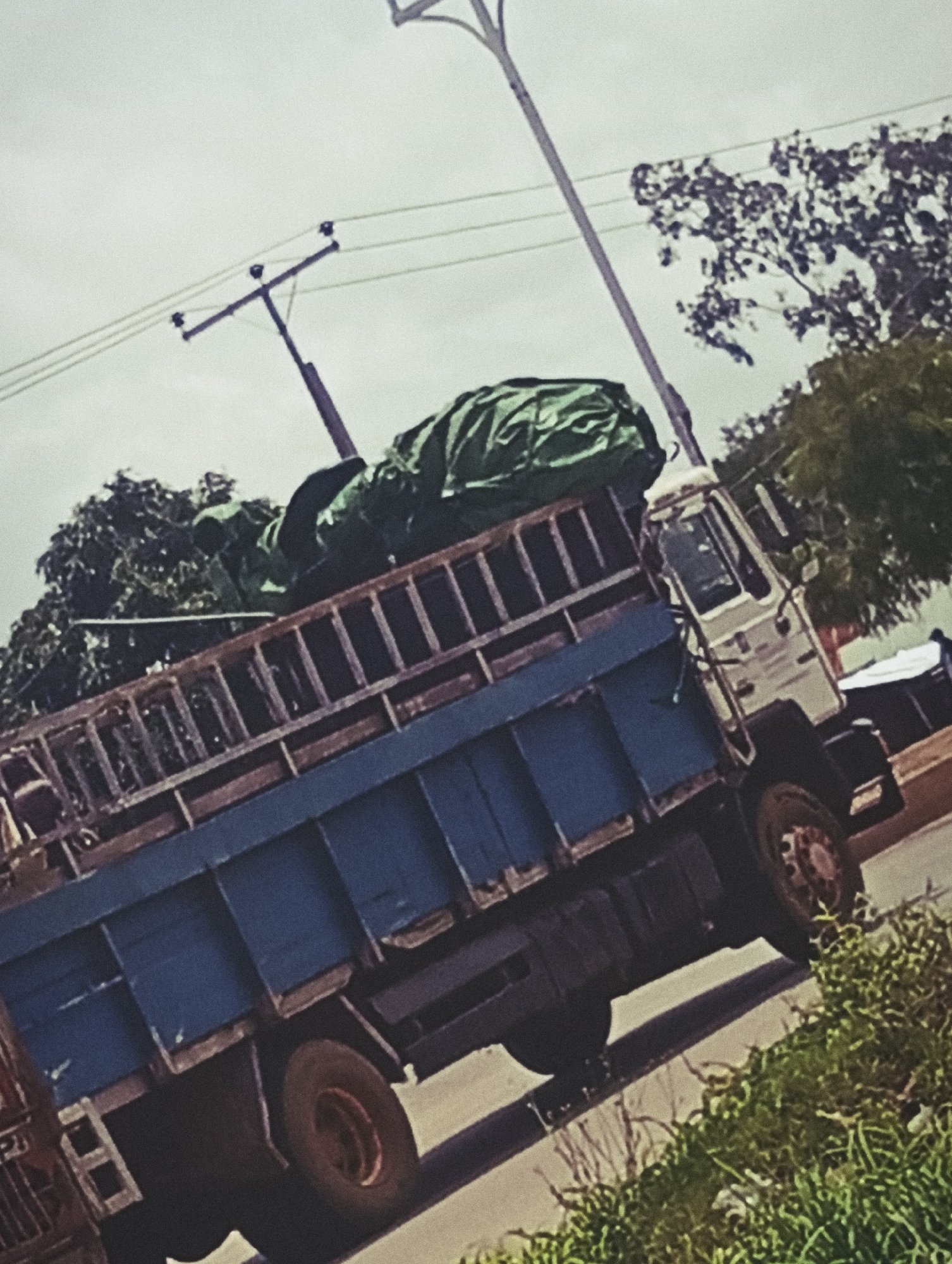 A tilted medium shot captures a blue flatbed truck loaded with green tarp-covered cargo. The truck's bed has wooden planks along the top edge. A white cab sits at the front, with a dark grille. Behind the truck, trees and vegetation are visible against a pale sky with power lines and a utility pole. A small portion of a white structure is to the right. The scene suggests a street in Bukuru, Nigeria, likely during daylight, given the ambient light and shadows. There are no visible people in the frame.