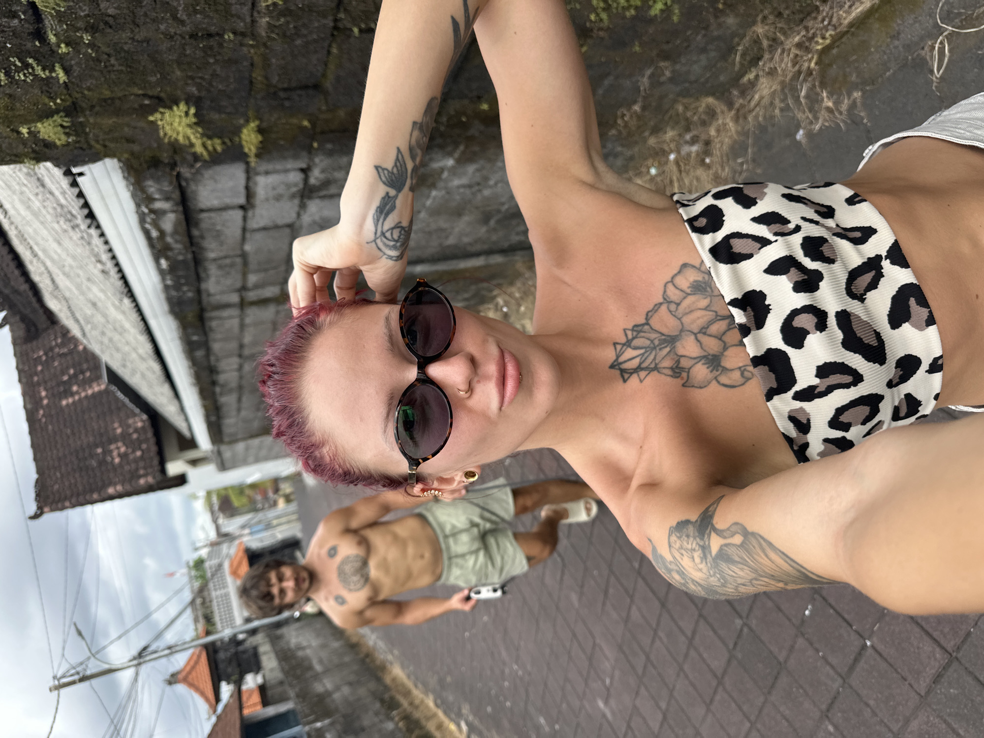 A low-angle medium shot depicts a person with short, reddish-purple hair, wearing sunglasses and a leopard-print top. They have multiple tattoos on their chest, arms, and hands. In the background, there is a shirtless person wearing green shorts and holding what appears to be a small, white electronic device. They also have visible tattoos on their chest. The setting seems to be a narrow street in Dalung, Indonesia. A tiled pathway is visible, with buildings and lush greenery lining the sides. The sky is overcast.