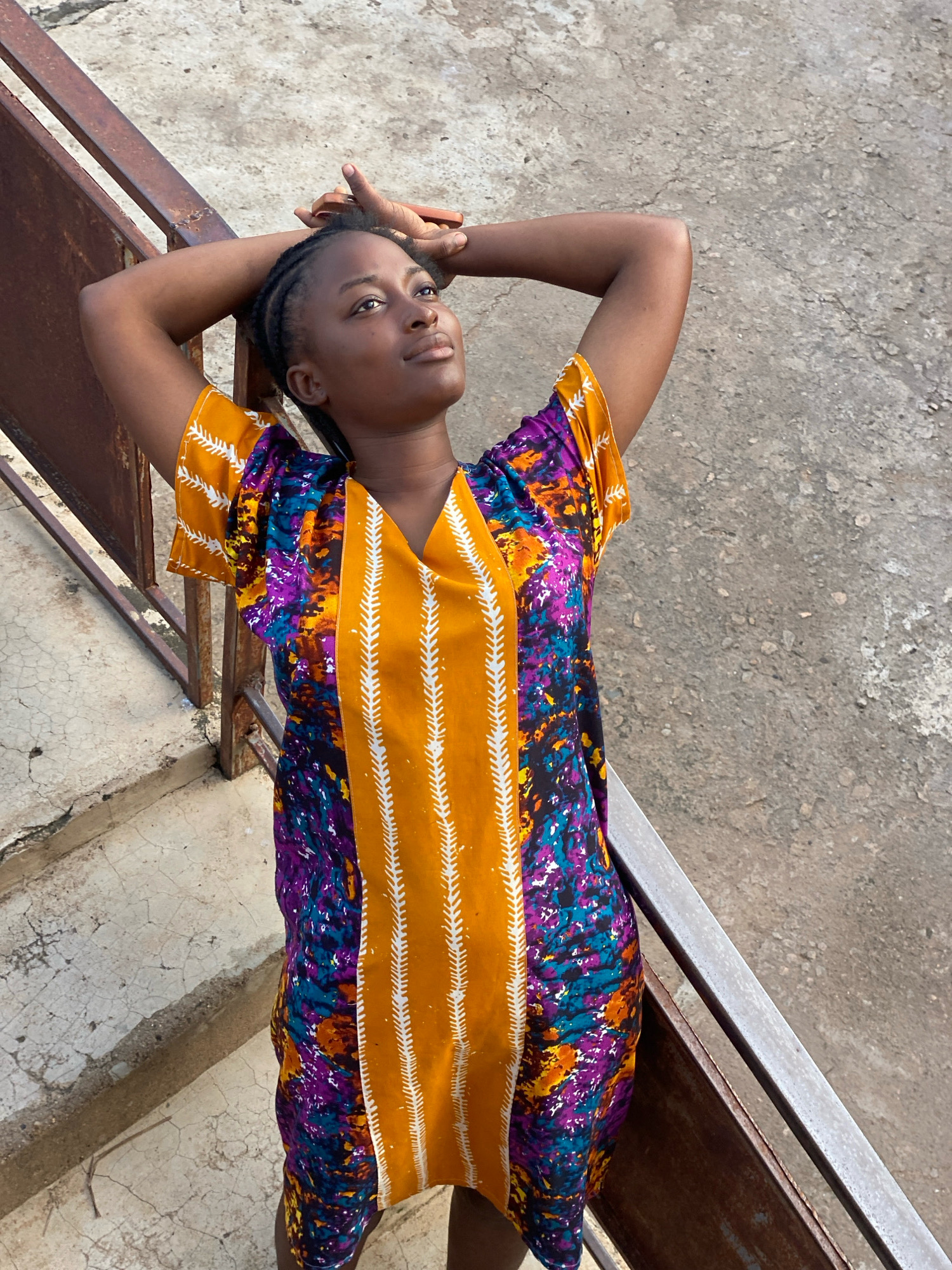 A young Black woman with braided hair stands on concrete stairs, her arms behind her head. She wears a brightly colored dress with an orange base and a purple and blue pattern. The stairs are worn and show signs of age, with a rusty metal railing on the left. The background is a textured concrete wall. The location is Sabon Tasha, Nigeria.
