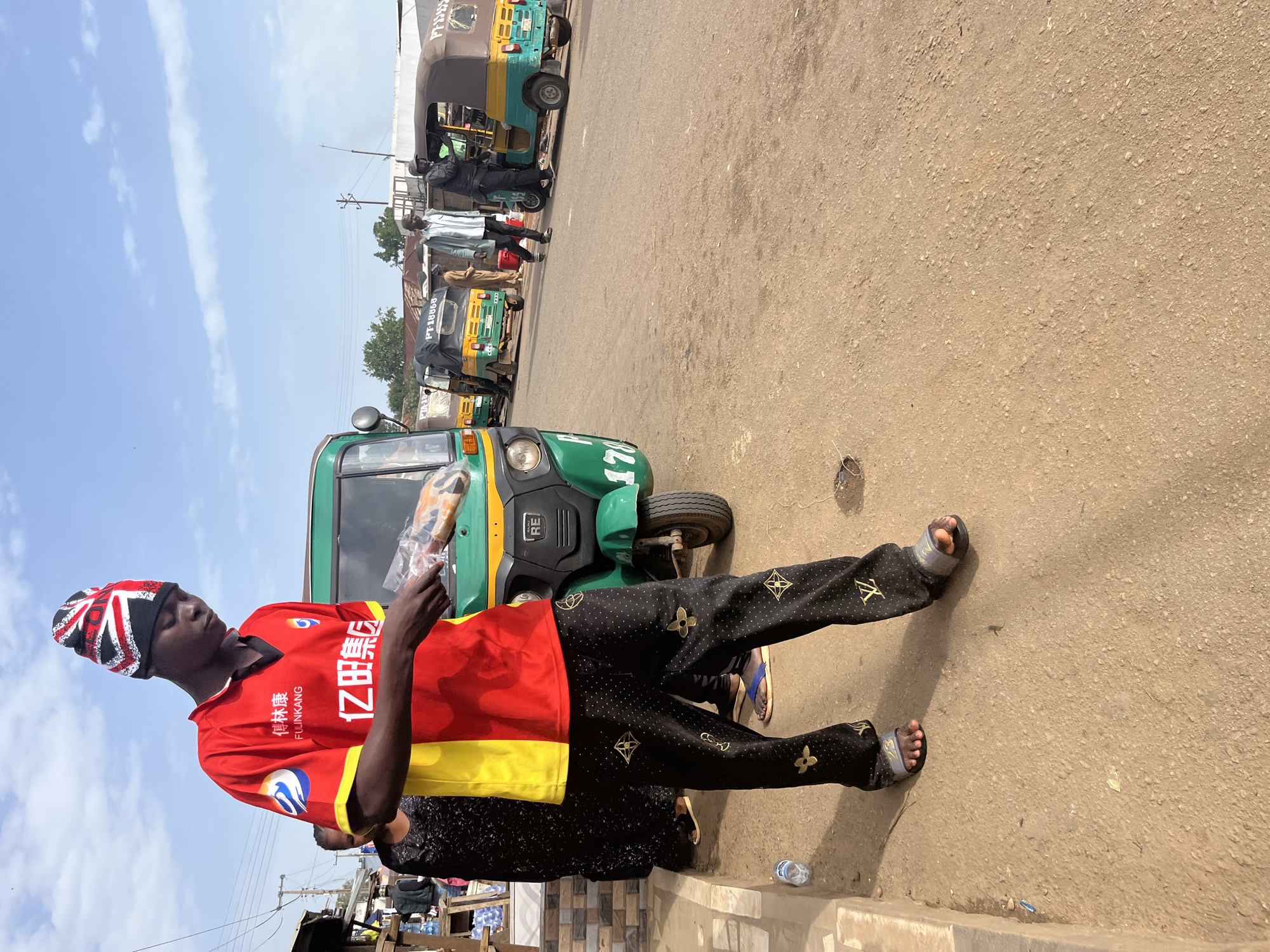 In Gura Top, Nigeria, a young Black male wearing a red and yellow shirt and black pants adorned with gold Louis Vuitton-style motifs stands on a dusty road. He wears a beanie with the Union Jack flag pattern. In his hands, he holds a clear plastic bag containing a long, light-brown object, possibly food. Behind him, a green and yellow auto-rickshaw is parked. Further down the road, other auto-rickshaws are visible, along with several Black individuals walking and standing. The environment appears to be an outdoor market or transportation hub under a bright, partly cloudy sky.