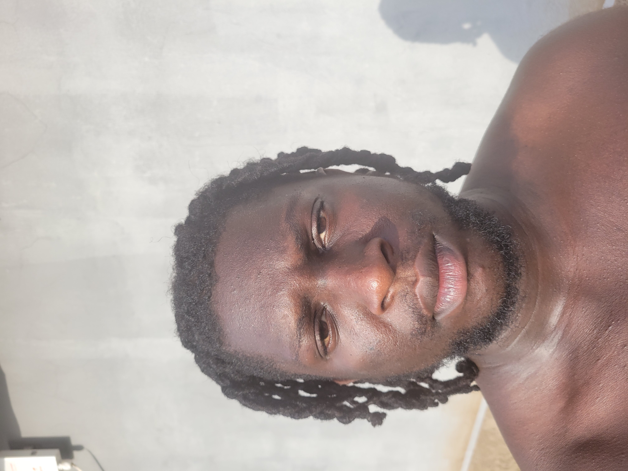 A close-up, angled portrait of a man with dark skin, dreadlocks, and a beard. He appears to be shirtless and is likely outdoors. The background is a light gray, possibly a concrete surface or wall. No specific details indicate the location as Markurdi, Nigeria.
