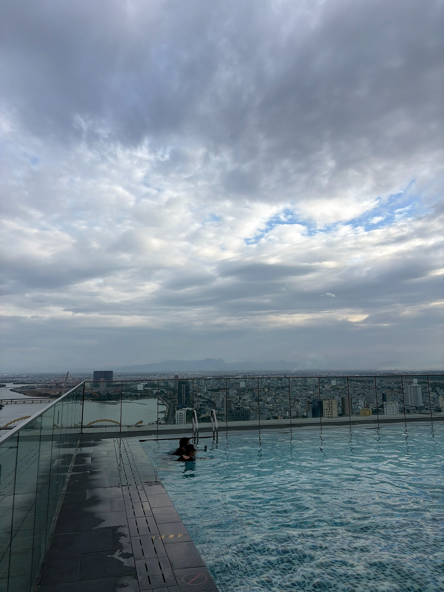 A person swims in a rooftop pool overlooking a city in Da Nang, Vietnam. The pool has a tiled bottom and is partially enclosed by a glass railing. The city is visible in the background, with a river, buildings, and mountains under a cloudy sky.