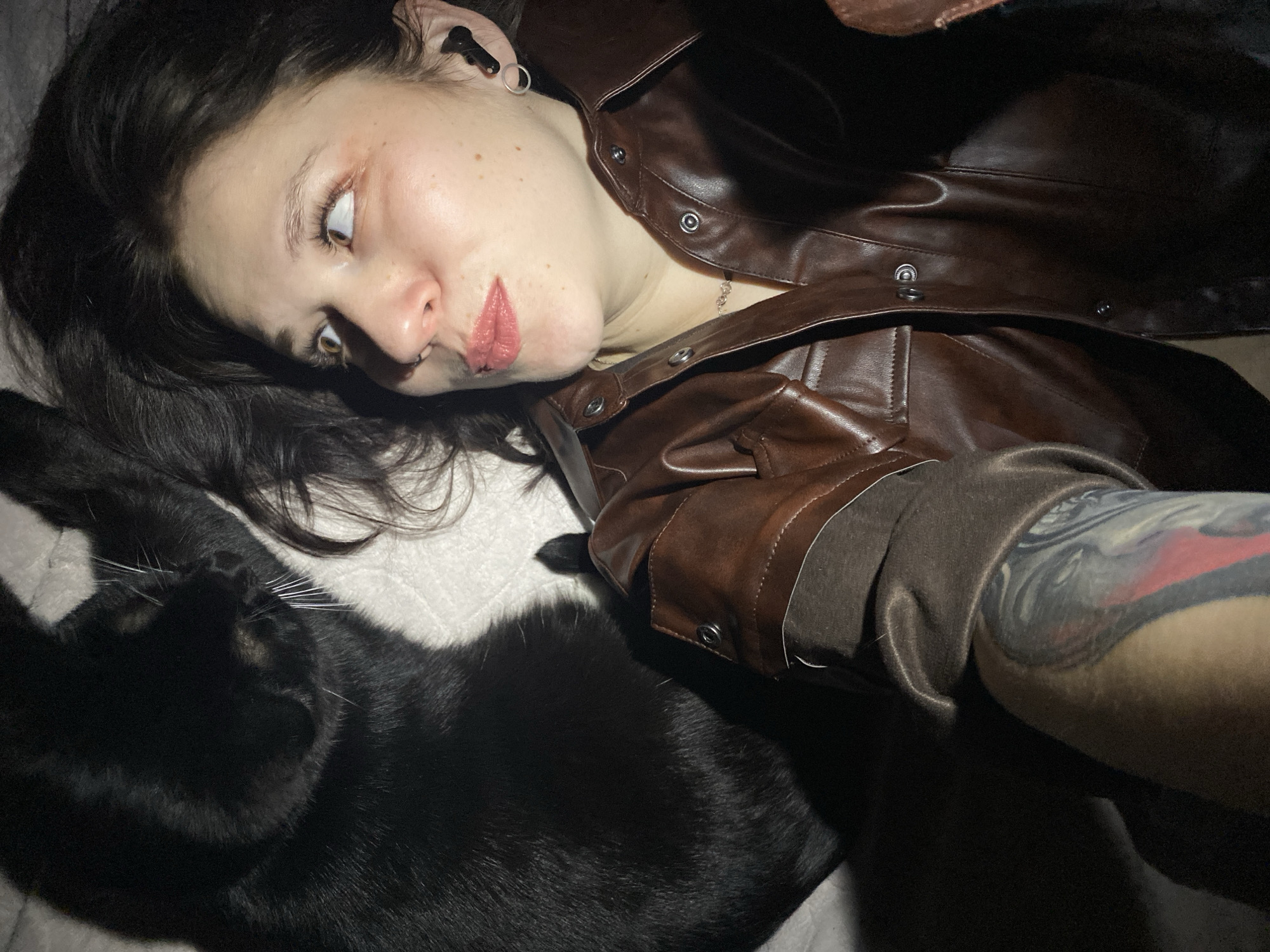 A person with fair skin and dark hair lies on a white surface, possibly a bed. The person is wearing a brown leather jacket and has a tattoo visible on their arm. A black cat rests beside the person. The background is dark, and the lighting is uneven, creating shadows. The city and country are not discernible from the provided image.