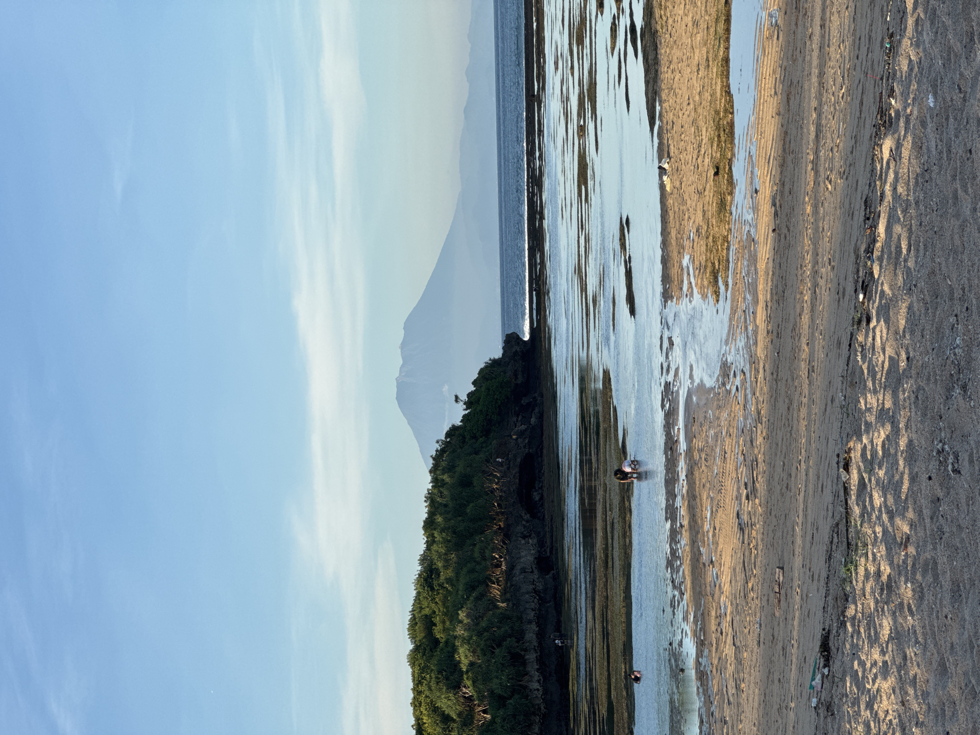 The image shows a beach scene in Kuta Selatan, Indonesia. The sky is a light blue with some wispy white clouds. To the left, there is an island covered in dense green vegetation, with a large mountain in the background. A body of water extends from the island to the right, covering parts of the sandy beach. Two people appear to be wading or searching in the shallow water. The sand on the beach varies in color and texture, with visible patterns from the water's edge. There is some debris scattered on the sand.
