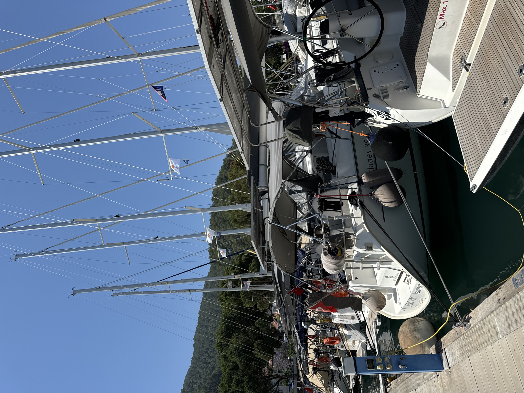 The image displays a marina scene with multiple sailboats docked along a pier under a clear blue sky, likely in the city of Fethiye, Turkey, given the prompt's context and the presence of Turkish flags. The bright sunlight suggests midday.
In the foreground, a sturdy pier is visible, constructed from wooden planks and metal grating. A blue utility pedestal equipped with several yellow and white power outlets stands on the pier. Yellow electrical cables extend from the pedestal to the docked boats.
A row of at least six sailboats is moored to the pier, facing away from the viewer. Most boats feature grey canvas biminis (awnings) covering their cockpits, and some have solar panels mounted on top of these awnings. Various boat accessories are visible, including grey and black fenders, orange life rings, and coiled ropes. On the stern of the sailboat closest to the left, the name "LAPIN" is clearly inscribed, along with "BENETEAU" above it, and the registration number "228758" and "KUSADASI" below. Another boat further right has "Elades 390" partially visible on its hull. The stern of the boat furthest to the right features a foldable swim platform with a graphic indicating "Max. 1" followed by a high-heel shoe symbol.
Several flags are flown from the masts of the sailboats. Turkish national flags (red with a white star and crescent) are visible on multiple vessels. Other flags include a white rectangular flag with an orange sun graphic and the number "203", and a dark blue pennant with white letters "OYK" and a red/pink symbol. A smaller, plain blue pennant is also visible.
In the background, steep, verdant hills covered with dense forest rise under the expanse of the cloudless blue sky. No people are visible in the scene.