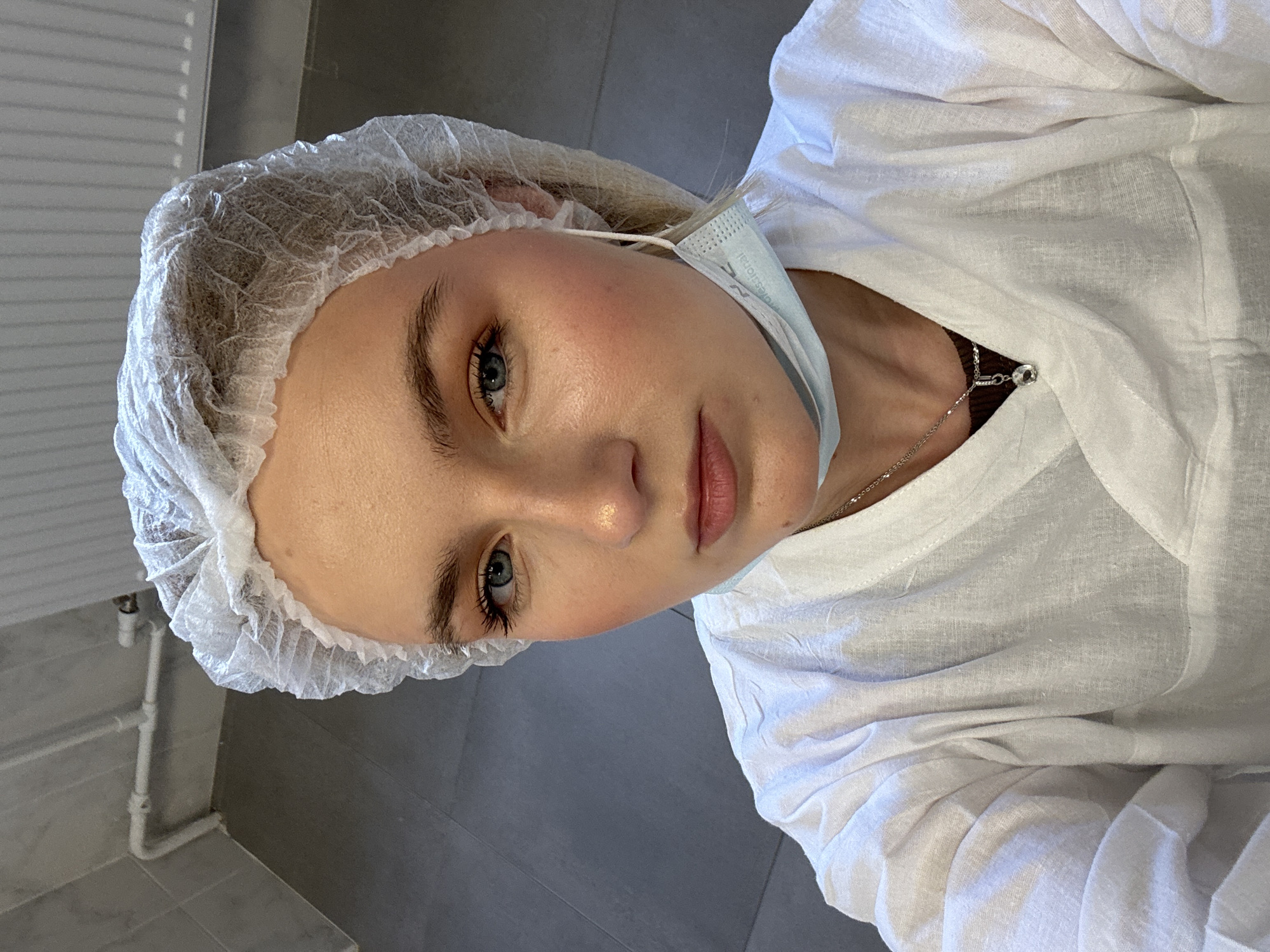 A woman is wearing a surgical cap, a white collared shirt, and a surgical mask hanging loosely around her neck. She has fair skin, blue eyes, and is wearing subtle makeup. A silver necklace is visible around her neck. The background consists of a tiled wall and what appears to be a radiator. The setting seems to be inside what could be a medical or cosmetic facility. It is not possible to determine if the image was taken in Saint Petersburg, Russia, based on the visual information provided.
