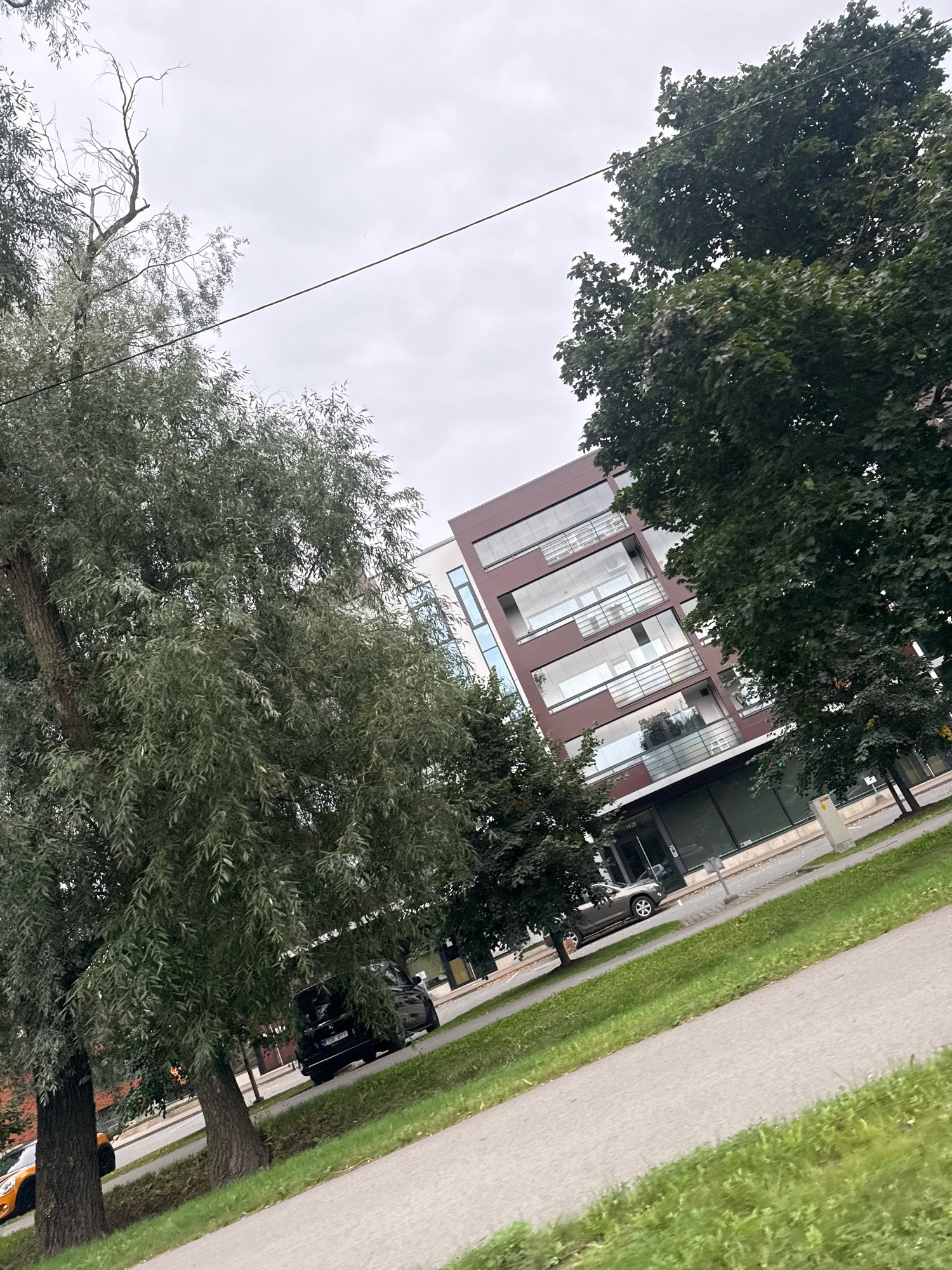 A modern, multi-story building with a dark brown facade and numerous balconies with glass railings is visible in Tallinn, Estonia. The building is situated along a street with a sidewalk and grassy areas. Trees with green foliage are in the foreground and partially obscure the building. Several cars are parked and driving on the street. The sky is overcast.