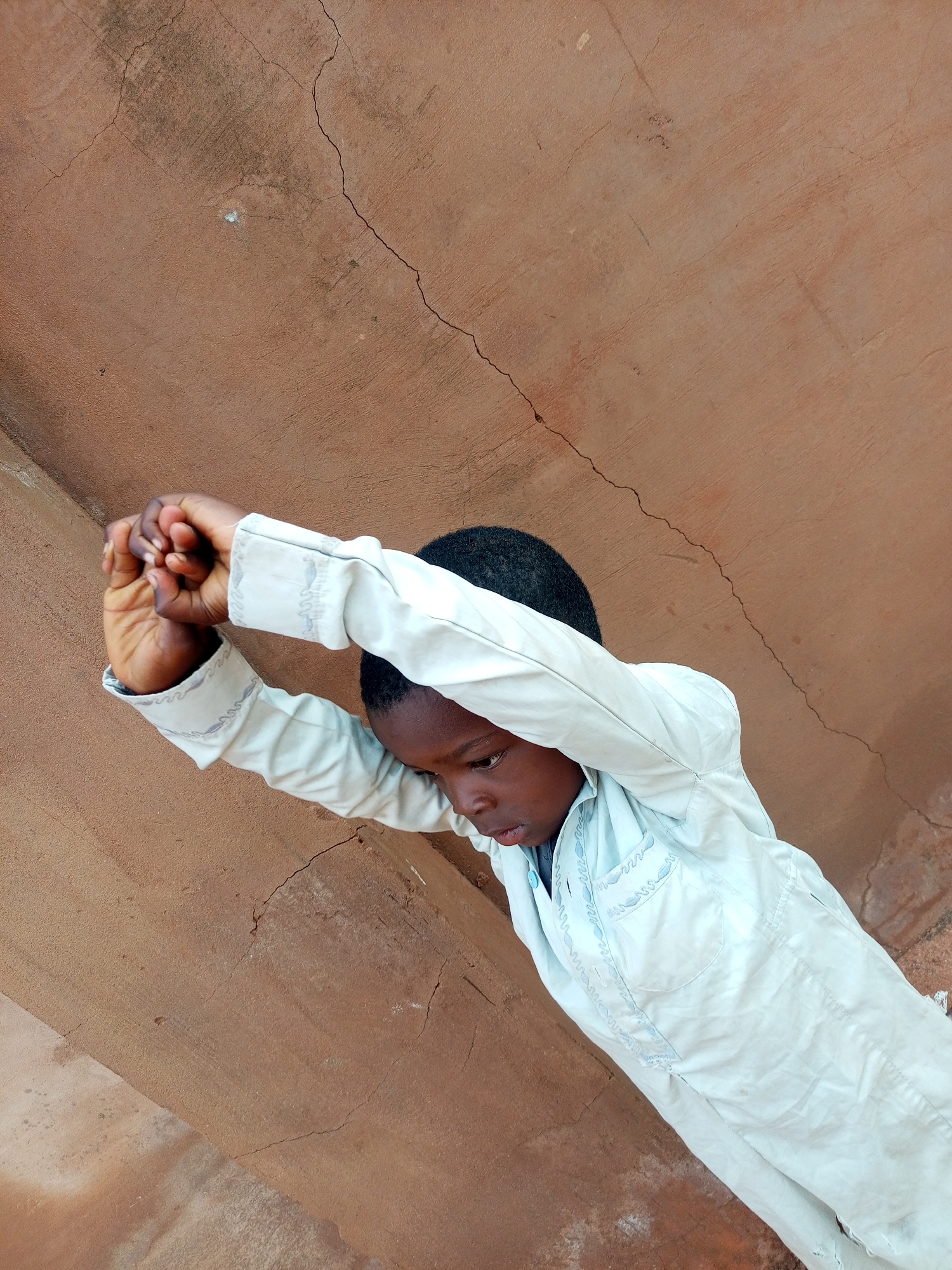 In Gombe, Nigeria, a young boy is captured in a downward-facing pose against a textured, reddish-brown wall. He is wearing a light blue garment with intricate white and light blue embroidery along the collar and pockets. His arms are raised and clasped together in front of him, his head is bowed, and his gaze is directed downwards. The wall behind him displays several cracks, indicating age or weathering. The lighting suggests an outdoor setting, possibly during the daytime.