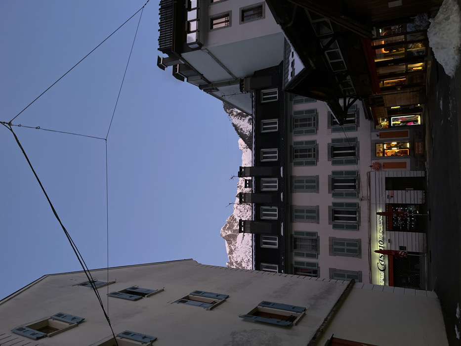 Buildings are vertically aligned, exhibiting a mix of white and dark facades. Shutters adorn the windows of a multi-story building. A nearby snow-capped mountain is visible. Overhead, a network of wires stretches across a clear sky. The foreground features a light-colored building with open windows. A store with lit windows and signage is prominent on the right.