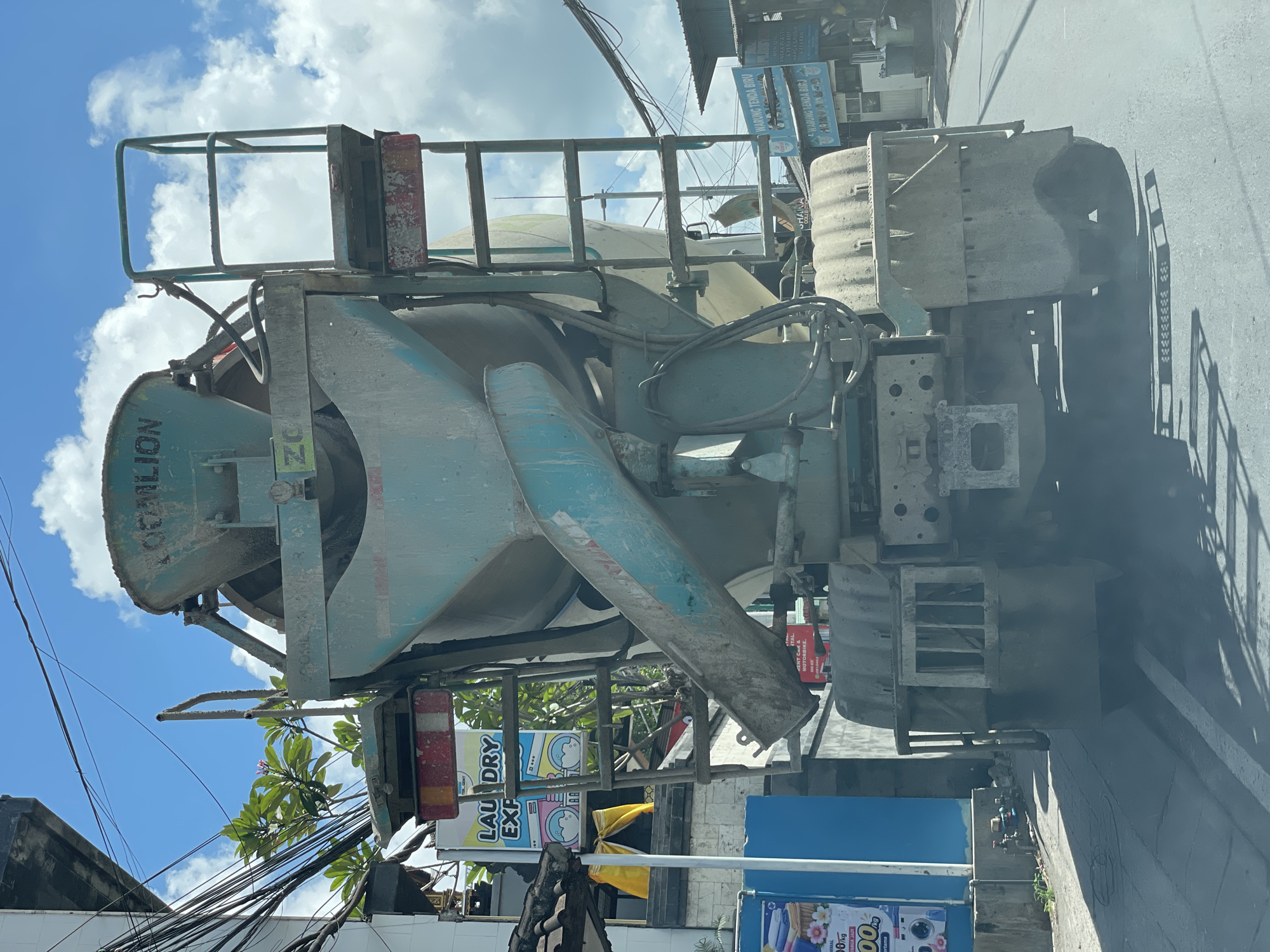 The image depicts the rear view of a cement mixer truck driving on a street in Kuta Selatan, Indonesia. The truck is light blue-green and appears dusty and weathered, with visible remnants of cement on its drum and chassis. The truck has a ladder on its left side leading up to a platform. The background reveals a street scene with various buildings, utility poles with a tangle of electrical wires, and lush green foliage. A sign for "LAUNDRY EXPRESS" with colorful cartoon images of washing machines is visible on the left. Further down the street, signs for "WARUNG TENDA BIRU" are present. The sky is bright blue with scattered white clouds, suggesting it is daytime. The ground is paved, and the truck's tires are visible on the asphalt. The overall atmosphere is that of a bustling, everyday street in a tropical urban environment.