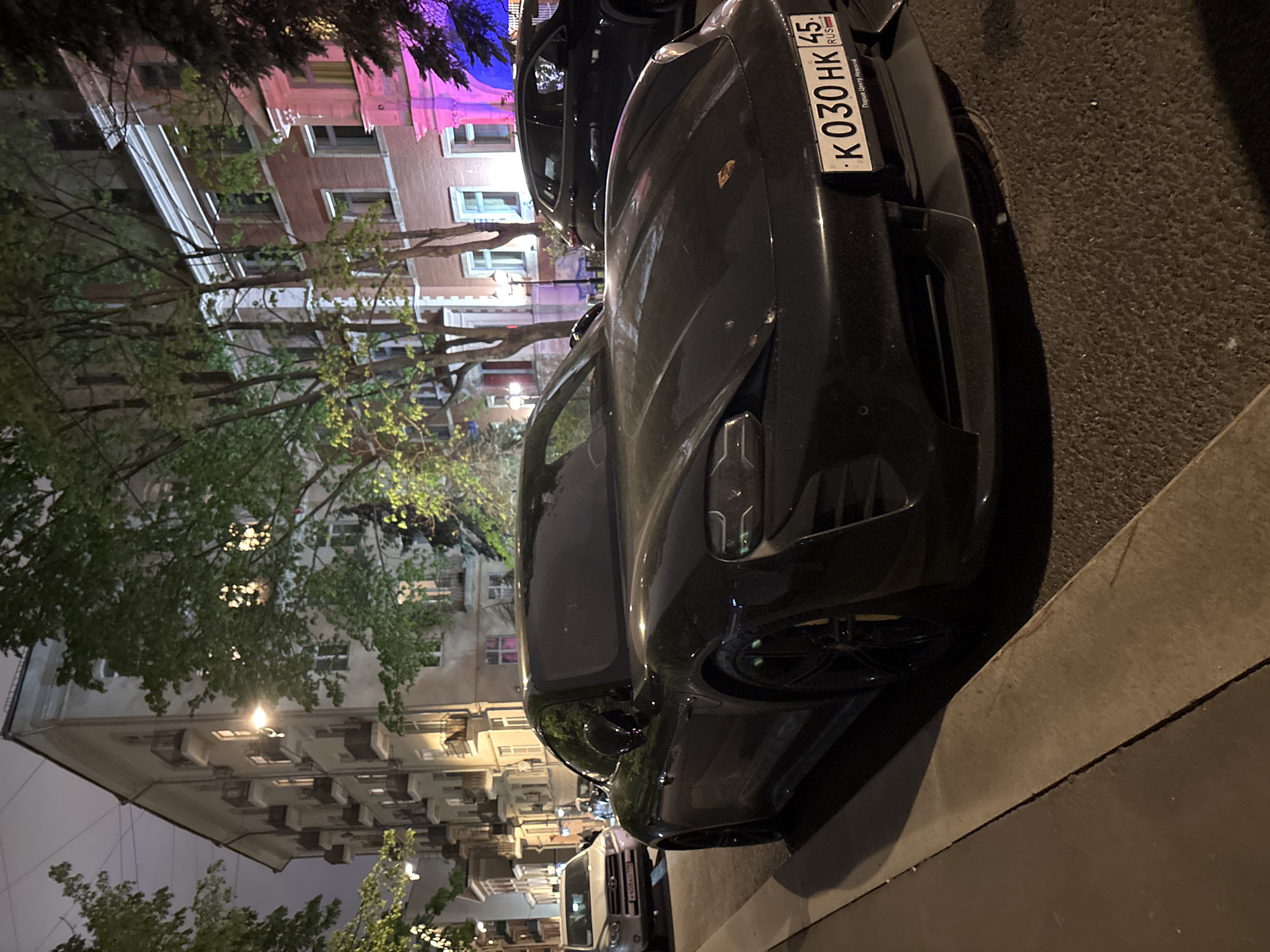 The photograph depicts a black Porsche parked on the side of a street in Moscow, Russia. The car has a Russian license plate that reads "K 030 HK 45 RUS." The background shows typical Moscow apartment buildings. There are trees along the street with their branches partially obscuring the buildings. The photo was taken at night under street lighting. There is another vehicle parked closely behind the Porsche.