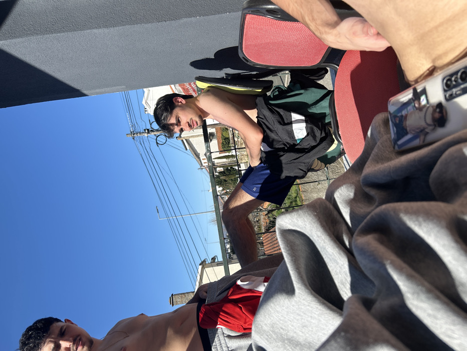 The image captures three individuals outdoors on a sunny day within a residential area of Bragança, Portugal.
In the left midground, a young male with dark hair and a mustache stands shirtless, wearing grey athletic shorts with a red garment partially visible inside. He faces the camera. A faint mark or scar is visible on his left chest.
To the right, a second young male with dark hair and a mustache is seated shirtless in a black office chair featuring a yellow-green backrest. He wears dark blue athletic shorts, and a black t-shirt with a white stripe is draped over the chair. He gazes towards the viewer's left. His left leg is extended forward.
In the immediate foreground, a substantial amount of grey fabric, consistent with clothing or a blanket, is positioned on the left. On the right, a white smartphone, encased in clear plastic displaying an inverted image of a person, rests on a red surface, likely the seat of a third chair. A partial view of a third individual's arm and leg is visible to the right of the smartphone.
The background features a clear blue sky. A metal railing separates the subjects from a residential panorama of houses with tiled roofs, one featuring a prominent stone chimney. Overhead utility lines, a street lamp, and sparse deciduous trees are present. A dark grey vertical structure on the far right casts a sharp shadow, indicating direct sunlight. A distant building displays a partial sign reading "eissell" or "elssele" followed by "serm" or "term". The setting appears to be a balcony or terrace.