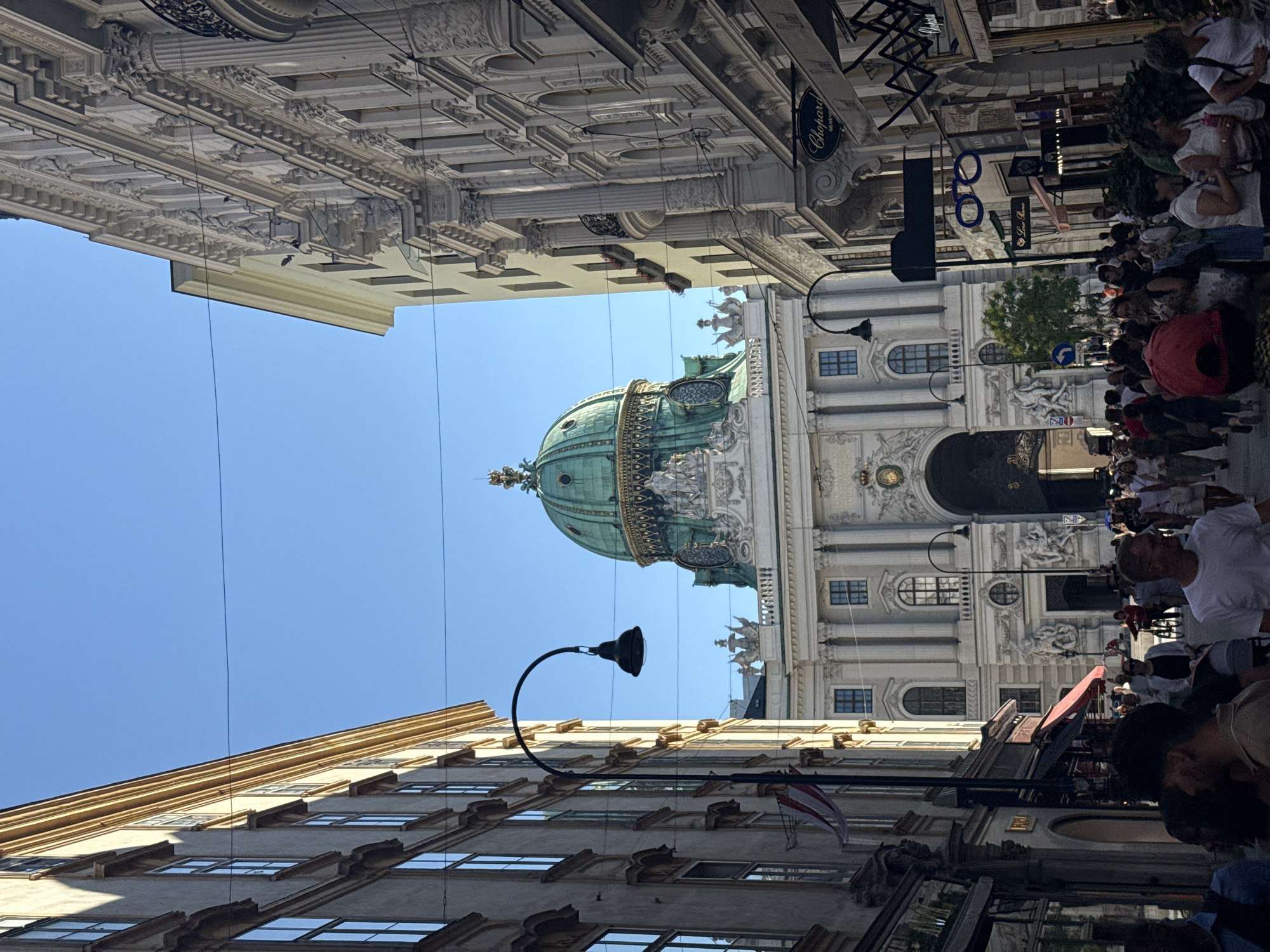 This is a media file capturing an upward-facing view of a city street in Vienna, Austria. The scene is dominated by ornate classical architecture. On the right, a crowd of people is visible, mostly walking or standing. Some are looking towards a grand building with a striking green dome, a prominent feature in the background. The dome is adorned with intricate details and sits atop a building with a light-colored facade, featuring multiple windows and decorative carvings. The sky is a clear, bright blue, suggesting a sunny day.
Street furniture such as lampposts and signs are also present. One lamppost, with a curved arm and a dark lamp, is particularly noticeable in the foreground. There are also various signs visible on the buildings, including a black sign with "Chopard GENEVE" written in white. The architectural style suggests a historic European city. The upward angle and the presence of people suggest a bustling street scene, possibly a tourist area. The overall impression is one of historical grandeur and a lively urban environment under a clear sky.