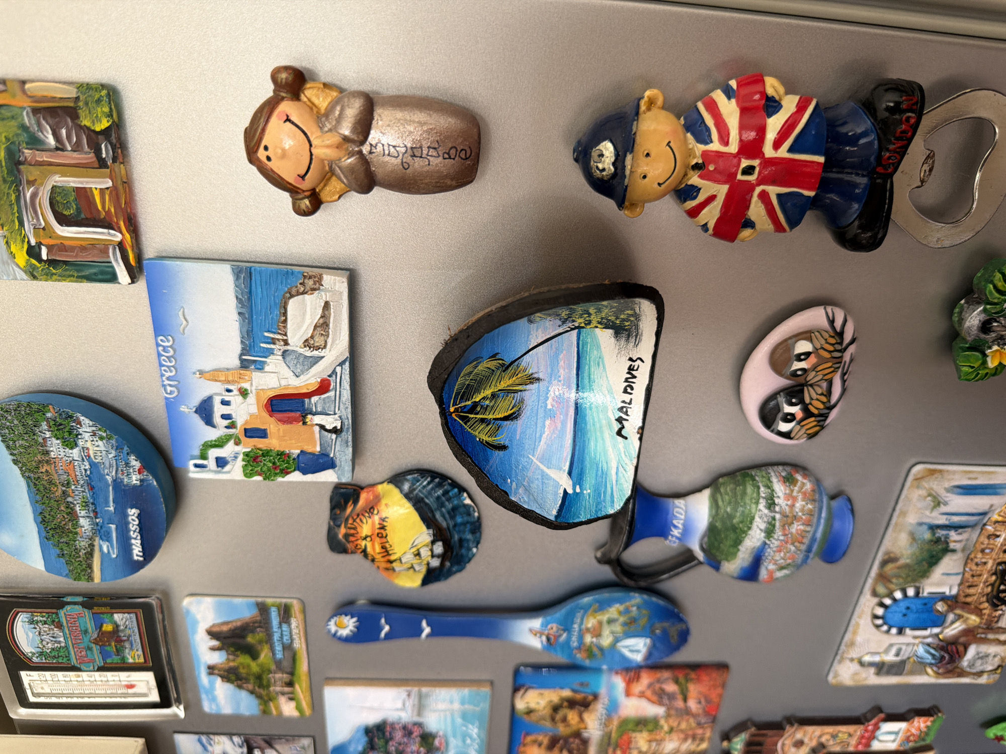 The image displays a collection of magnets on a metallic surface, likely a refrigerator door. The magnets feature various tourist attractions and souvenirs from different locations. Notable magnets include a thermometer magnet for "West Virginia," a round magnet depicting "THASSOS," a rectangular magnet with a Greek village scene labeled "Greece," and a shell-shaped magnet with a ship and the text "SAINT HELENA." Other magnets show a palm tree beach scene with "MALDIVES" written on it, a blue spoon magnet with various illustrations, a jug magnet with a landscape and the text "FKADA," a small oval magnet with two owls, and a magnet depicting a camel and a blue doorway. There is also a figurine of a person in a police uniform wearing a Union Jack-themed coat, with "LONDON" written on their shoes, which also functions as a bottle opener. Another magnet is a figurine of a person with their hands clasped in prayer. The overall environment is cluttered with these decorative magnets. The city of Pazardzhik, Bulgaria is not depicted in this image.