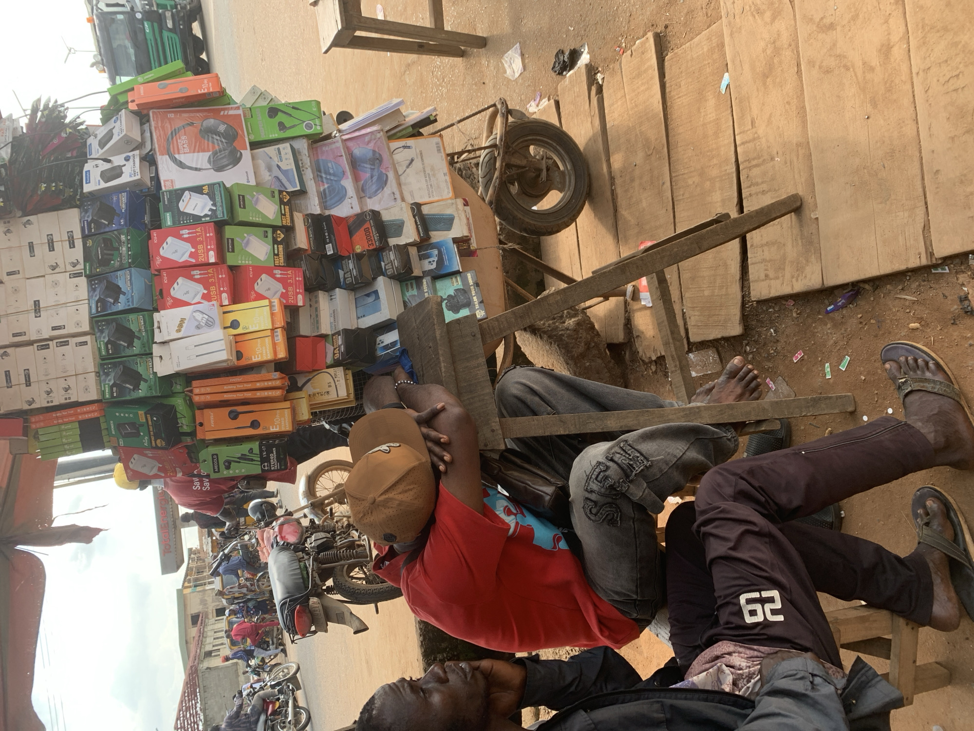 This media file captures a busy street scene in Shagamu, Nigeria, likely during the daytime, given the bright lighting. The foreground is dominated by a man wearing a red t-shirt, a beige baseball cap, and jeans, resting on a makeshift wooden structure. He appears relaxed, with his arm resting on his head and a brown bag beside him. Another person, wearing dark pants with the number "62" prominently displayed, is also seated.
Behind the seated individuals, a market stall is packed with an assortment of electronics, primarily headphones and chargers in colorful packaging. Prominent text includes brand names like "JBL," "AREGOLD," and "Total Energies." The stall is built on a cart with a large wheel, suggesting it might be mobile.
In the background, a street is filled with various motorcycles and a few other individuals. The general atmosphere suggests a bustling commercial area. The presence of a large truck on the left side of the image indicates ongoing traffic. The scene conveys a sense of everyday life and commerce in a Nigerian urban environment.