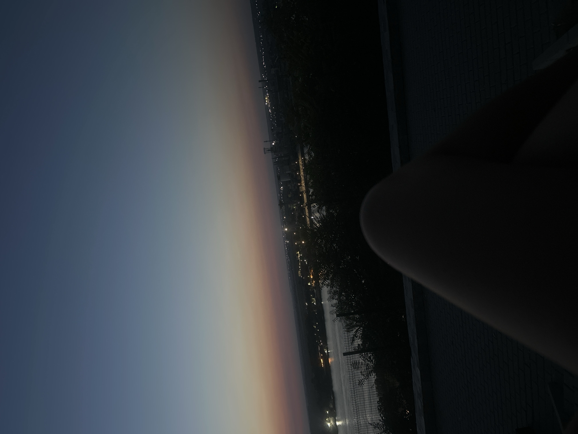 The image depicts a dusky twilight scene overlooking a city and a body of water, likely the Danube River, in Nikopol, Bulgaria. In the foreground, a dark, out-of-focus shape, possibly a person's leg, occupies the lower portion of the frame. Beyond this, a dark, tiled surface stretches towards the middle ground. A low fence runs horizontally across the mid-section of the image. Across the water, the city lights of Nikopol twinkle, with industrial structures and buildings visible in the distance. The sky exhibits a gradient of deep blue at the top, transitioning through shades of purple and pink near the horizon, indicating the end of daylight. The overall impression is one of a serene, urban landscape at dusk.