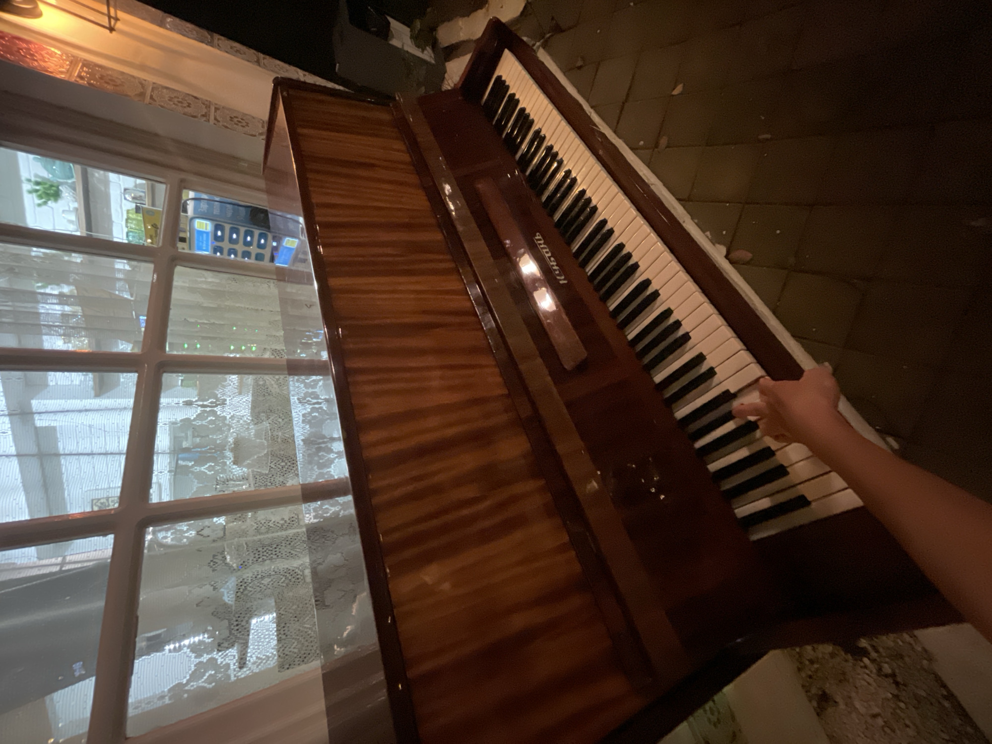 An upright piano with a polished wood finish is positioned in front of a large window with sheer curtains. A hand is reaching towards the keys, suggesting someone is about to play. Through the window, a shelf with decorative items and a mobile phone charging on a device can be seen. The floor beneath the piano is tiled. The scene appears to be indoors, possibly in a residential setting in Sochi, Russia.