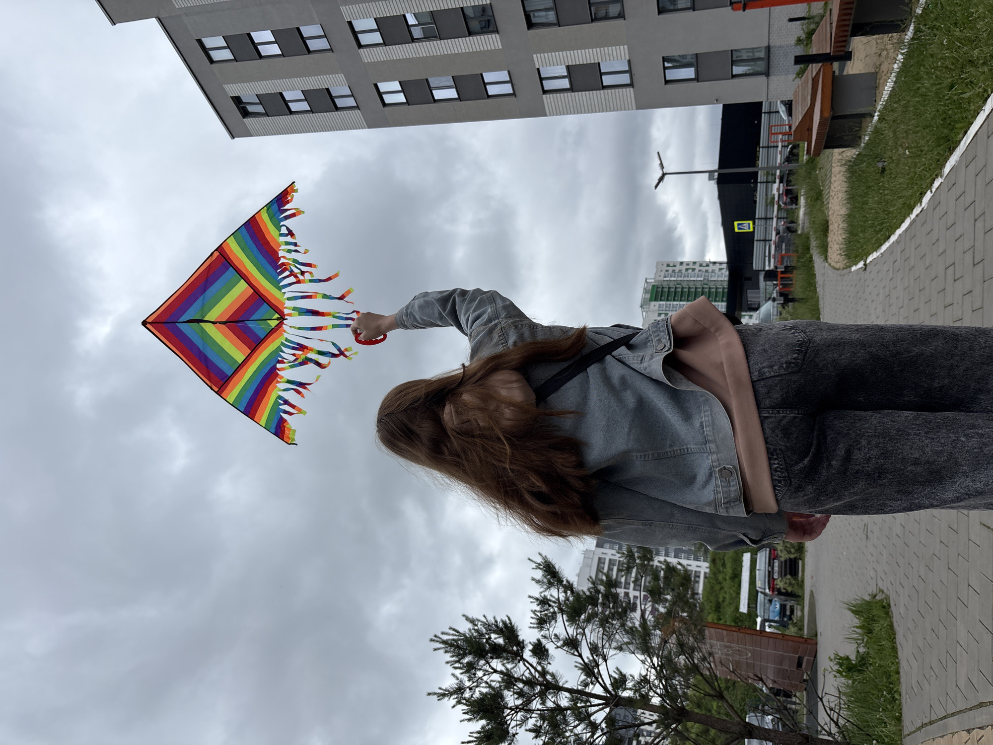 An outdoor, eye-level, full shot shows a person standing on a paved walkway, viewed from the back. The person has long, light brown hair and is wearing a light blue denim jacket over a peach-colored top and dark grey jeans. Their right arm is raised, holding a large, square, rainbow-striped kite with a red plastic handle. The kite features colorful streamers cascading from its lower edge against an overcast sky filled with grey clouds. To the left of the person, a sparse pine tree stands beside the paved path, with a white, multi-story residential building visible behind it, and a distant building with green architectural elements further left. To the right, a tall, modern, grey apartment building with dark-framed windows and light grey vertical textured panels occupies the background. A black street lamp with a single luminaire is positioned in front of this building. The paved walkway, constructed from light grey rectangular bricks, curves slightly to the right, bordered by a strip of grass and two wooden benches. In the far background, a dark commercial building with a yellow pedestrian crossing sign is discernible. The scene depicts an urban landscape in Izhevsk, Russia.