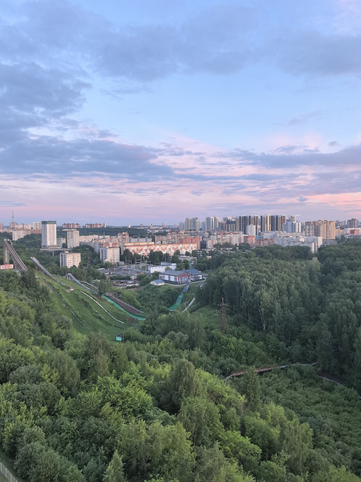 The image depicts a panoramic view of the city of Perm, Russia, at what appears to be dawn or dusk, given the pink hues in the sky. The foreground is dominated by a dense green forest, with trees varying in height and density. Winding paths are visible cutting through the trees.
In the middle ground, there are distinct ski jump facilities, including structures for launch and landing. The ski slopes are visible as grassy, sloped areas that lead down through the trees.
Beyond the forest and ski jumps is the city itself. It is characterized by a mix of low-rise and high-rise buildings, typical of urban development. The buildings are primarily residential. In the background, more high-rise buildings are distinguishable, indicating a dense urban center.
The sky is a mix of blue and pink, indicating either sunrise or sunset. Clouds are scattered across the sky.