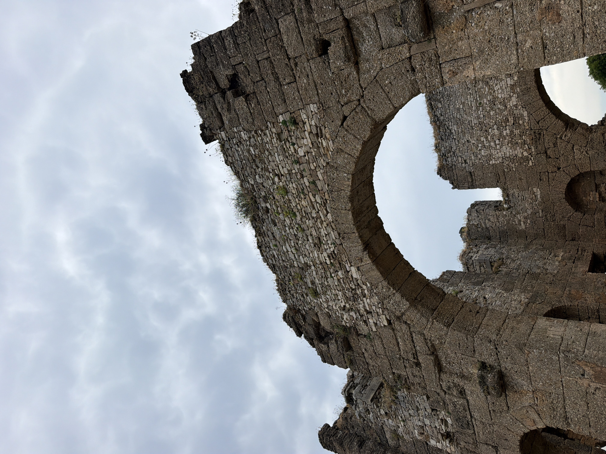 The image shows an arched stone structure, likely part of ancient ruins, with a cloudy sky in the background. The stonework appears weathered and aged, with some smaller stones filling gaps in larger blocks. Vegetation, including small plants and grasses, grows from crevices in the stone. The structure is made of large, rough-cut stones in its lower sections and smaller, more regular stones in the upper portions and arch. The perspective is looking upwards towards the arch and the sky. This scene is characteristic of the ancient ruins found in and around Serik, Turkey, such as those in the Aspendos theater or ancient city.