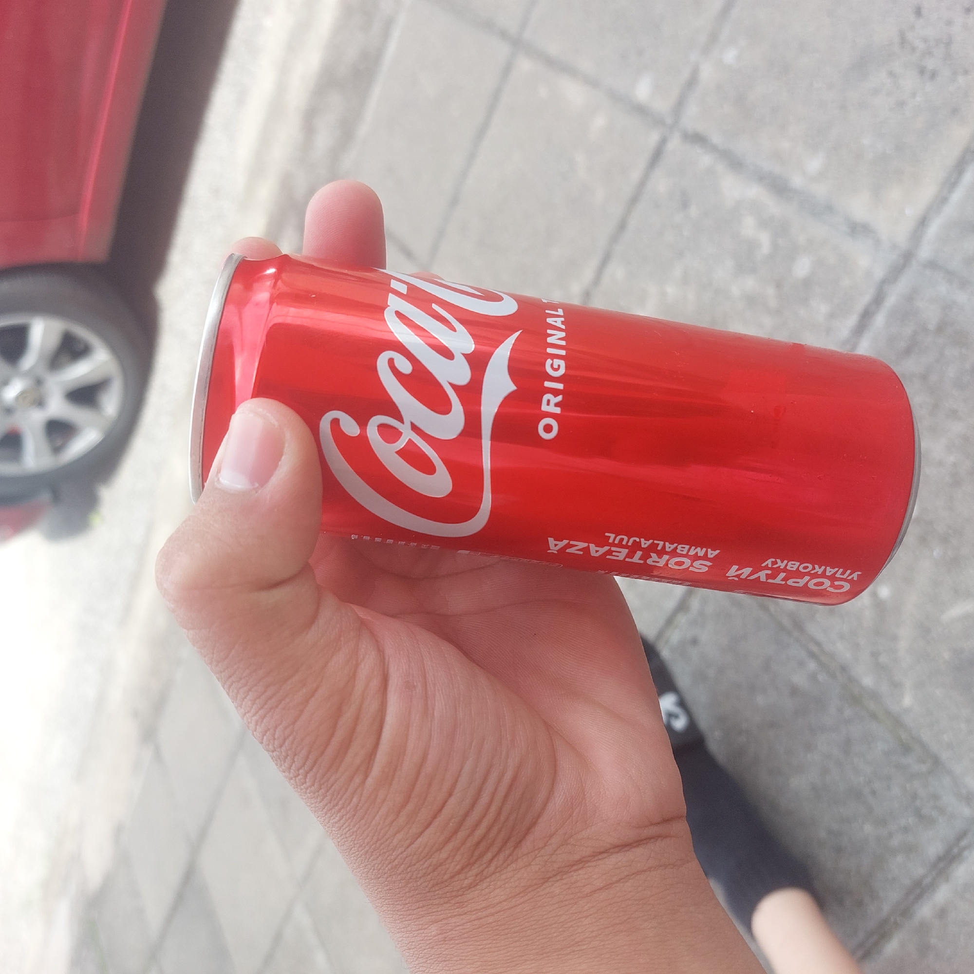 A person is holding a red Coca-Cola can in their right hand. The can has the Coca-Cola logo in white script and the word "ORIGINAL" below it. There are also some vertical lines of text in a foreign language on the left side of the can. The background is a blurred outdoor scene with a paved surface and a red car. The image appears to be taken in Ablanitsa, Bulgaria.