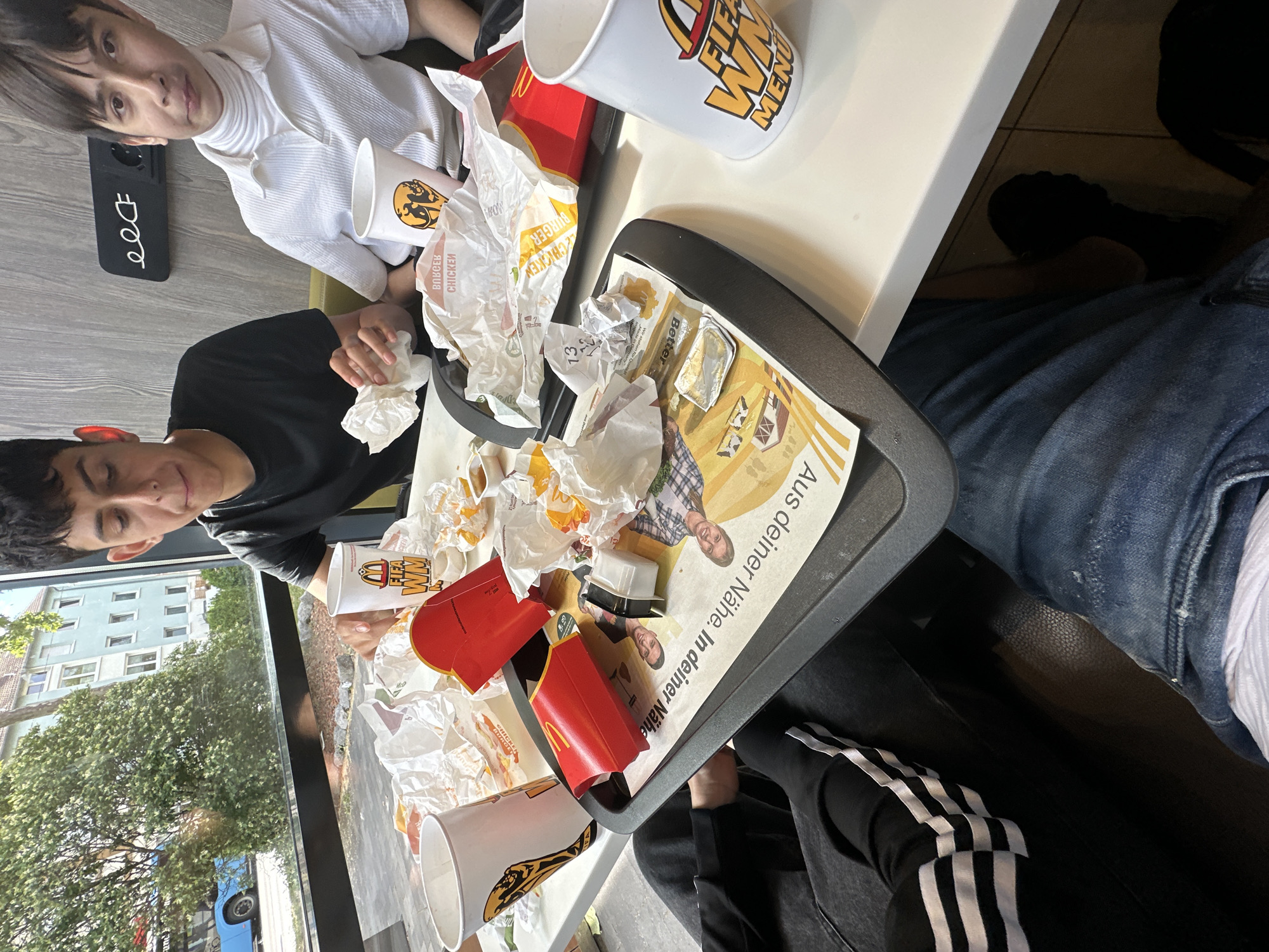 The image depicts a table with fast food remnants and two young males. A dark-haired male in a black t-shirt is seated on the left, holding a white napkin. To his right, a male with dark hair and a white turtleneck shirt is visible. Both are seated at a table with used McDonald's packaging, cups, and wrappers. The table is set against a backdrop of a large window overlooking a street with a blue bus and buildings. A black sign with a white plug symbol is mounted on the wall behind the individuals. The scene appears to be a casual dining setting.