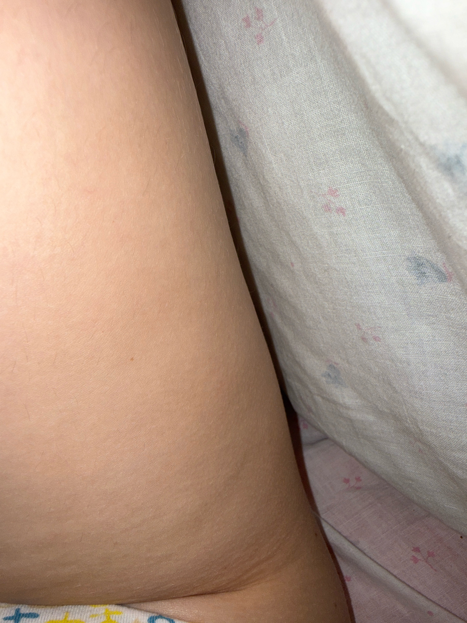 The image is a close-up, eye-level shot of a person's leg and a patterned cloth, possibly a blanket or sheet. The leg is smooth and lightly tanned, with a few faint hairs visible. A small, dark mole is present on the skin. To the right of the leg, the cloth is draped and folded, revealing a pale pink and white fabric with a subtle pattern of small, scattered pink and blue shapes that resemble flowers or paw prints. The lighting is soft, creating gentle shadows on the skin and fabric. The overall impression is one of intimacy and domesticity, likely taken in a bedroom. There is no information about the location in Zavolzhye, Russia, visible in the image.