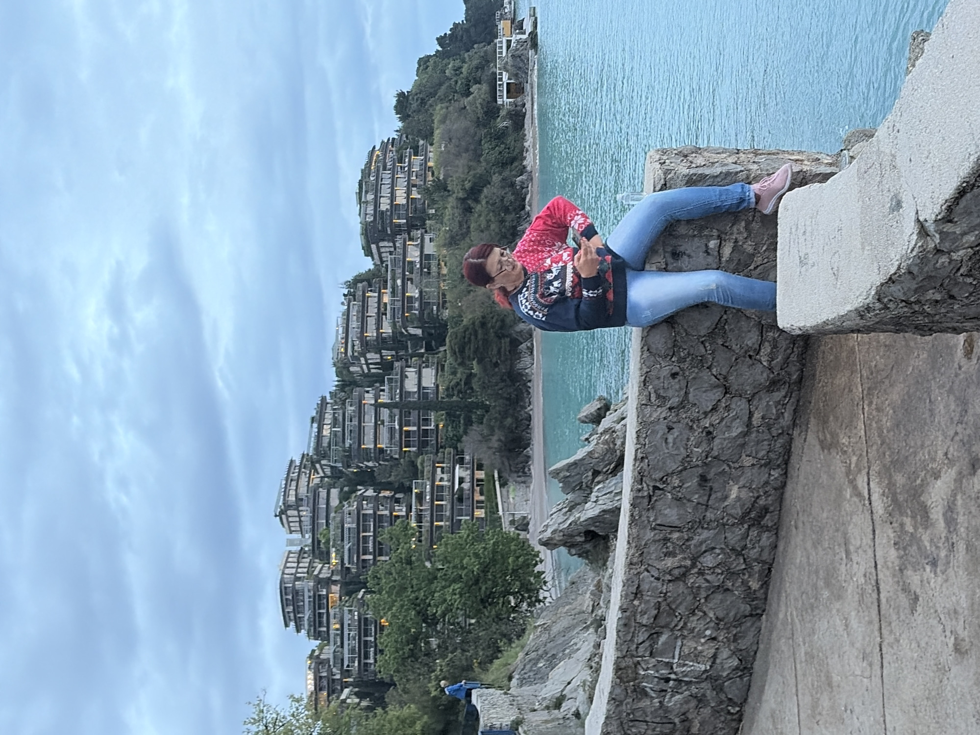 A photograph taken in Budva, Montenegro, depicts a woman with red hair wearing a red and blue patterned sweater and blue jeans, sitting on a stone ledge with her legs extended towards the ocean. She is looking at the camera and making a hand gesture. The ocean, visible in the background, is a clear turquoise color. Behind the woman and the ocean are tiered apartment buildings that line the coastline. The sky is cloudy. In the foreground, the stone ledge she is sitting on is textured.