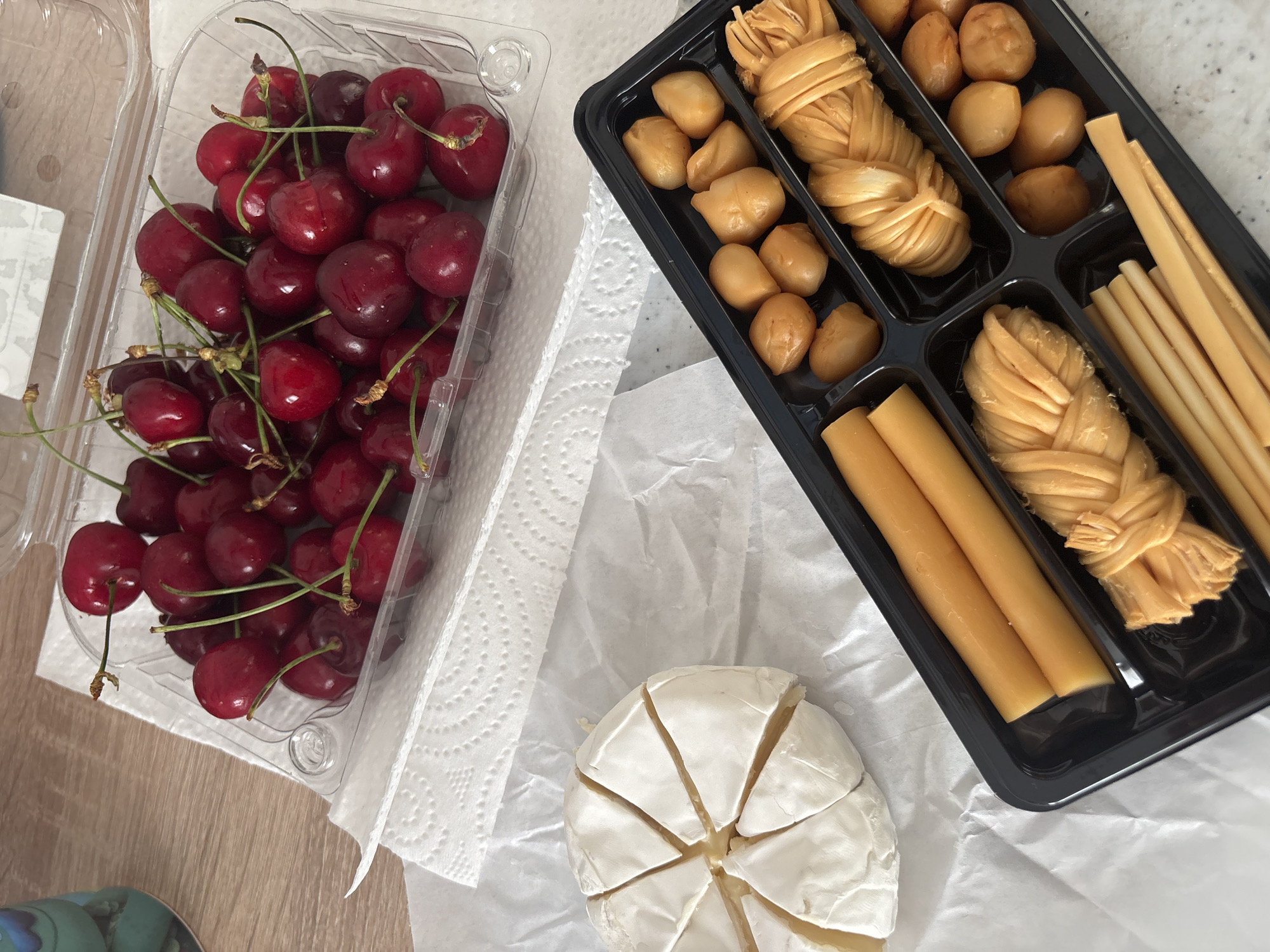 The image provides an overhead view of various food items arranged on a tabletop. On the left, a clear plastic container holds a generous quantity of fresh, deep red cherries, most still attached to their green stems. To the right of the cherries, a black plastic tray is compartmentalized, filled with an assortment of cheese snacks. These include numerous small, spherical or oval-shaped, golden-brown pieces, as well as distinct portions of yellowish, intricately braided or woven cheese. Another compartment contains several straight, cylindrical cheese sticks or rods.
Below the black tray, a round wheel of soft white cheese, resembling Brie or Camembert, is visibly pre-cut into several triangular wedges and rests on a sheet of white parchment paper. White paper towels are spread beneath the food items on the table, which has a light wooden surface partially visible on the left. A small segment of a blue and green object, possibly a cup or mug, can be seen at the very bottom-left corner of the frame. The scene appears to be indoors, under bright, possibly natural, lighting. No people or visible text are present in the image.