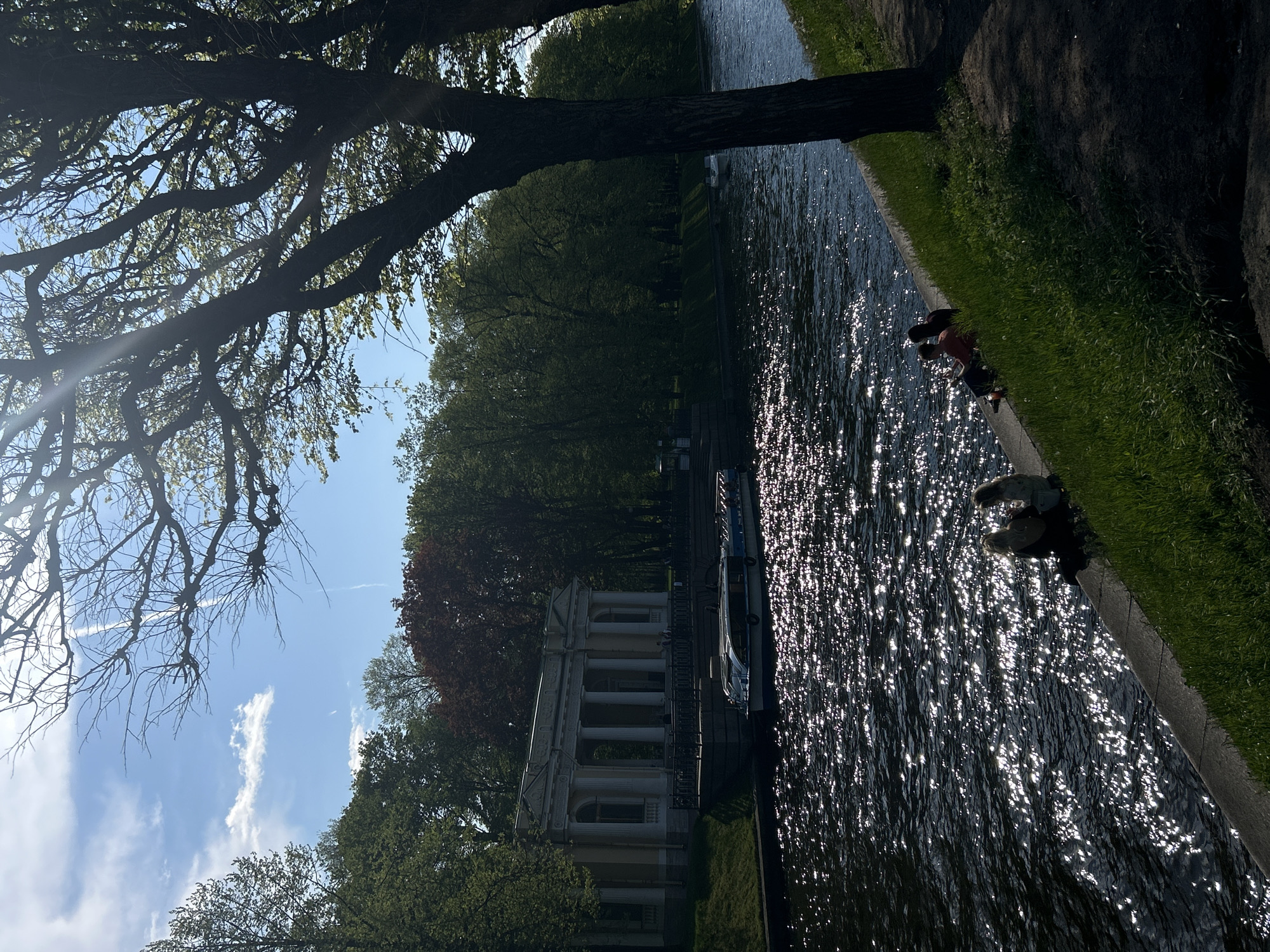 The image depicts a canal in Saint Petersburg, Russia. The view is partially obstructed by a large tree branch in the upper-left corner. On the left side of the canal, a classical building with columns is visible. A boat is moored alongside the canal. On the right bank, two people are interacting near the edge of the canal. The water reflects sunlight, creating bright spots on the surface. The sky is blue with scattered clouds.