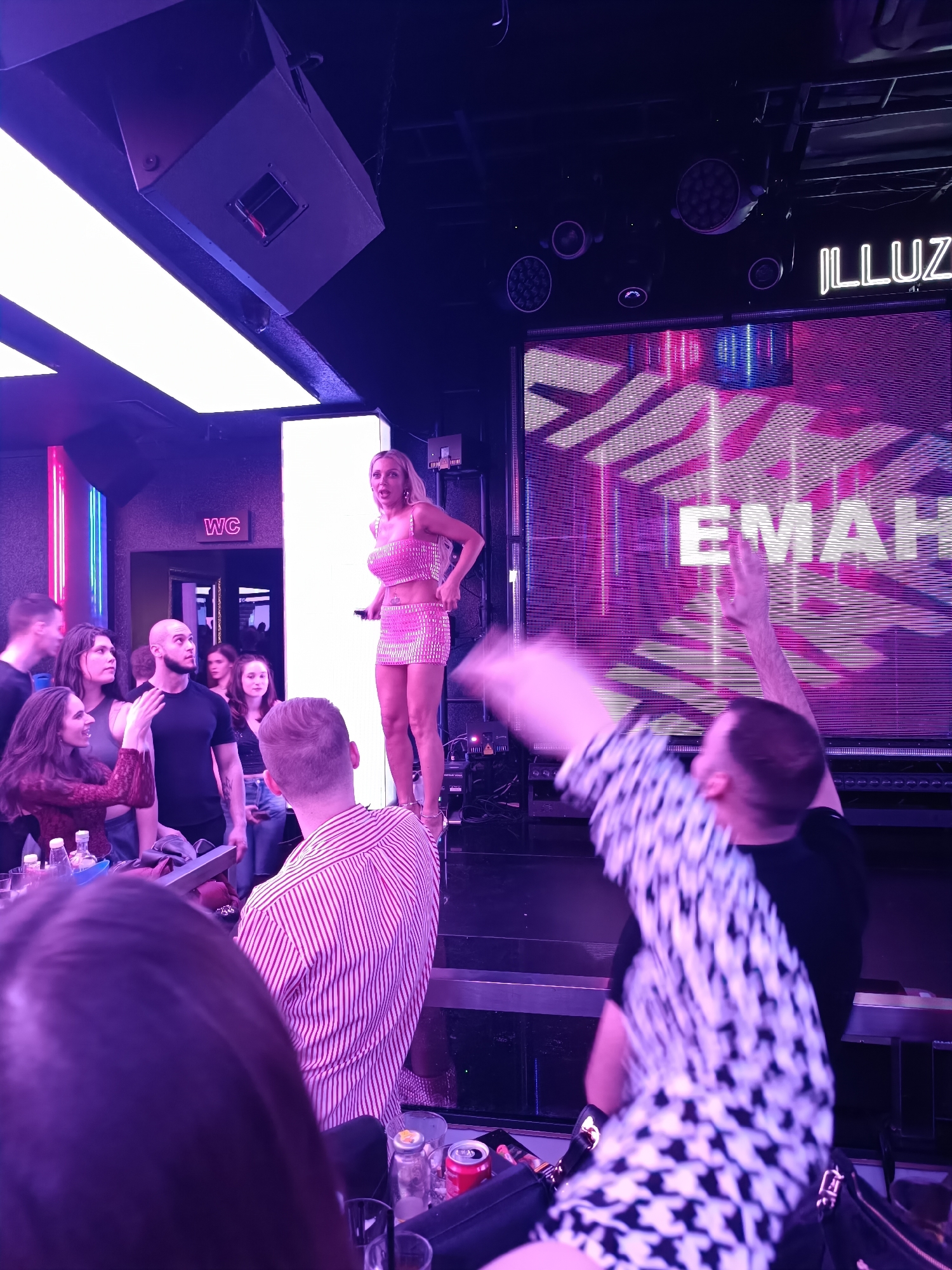 An indoor scene depicts a lively nightclub, identified as ILLUZION Club in Sofia, Bulgaria, by neon signage reading "ILLUZ" in the upper right. The environment is illuminated predominantly by purple and blue lighting. A blonde female performer, wearing a two-piece sparkly pink crop top and mini skirt with high heels, stands on a central elevated platform. She holds a microphone and her mouth is open, suggesting she is singing or speaking.
She is flanked by two tall, vertical white light panels. Behind her and to her right, a large horizontal LED screen displays dynamic purple and blue geometric patterns and the white text "EMAH". Overhead, various sound speakers and lighting fixtures, including spotlights and linear white lights, are visible against a dark ceiling.
Below the stage, a crowd of approximately ten individuals is observing. A bald man in a black t-shirt looks directly up at the performer. To his left, a woman in a reddish top gestures with her hand while appearing to engage in conversation with the bald man. In the foreground, from a higher vantage point, the back of a person with dark hair is visible. A dark surface in the foreground displays bottles, glasses, and a red Coca-Cola can. To the right, another individual, viewed from the back and wearing a black and white patterned shirt, has both arms raised in a blurred motion, indicating active participation or dancing. A "WC" sign is visible on a door in the background.