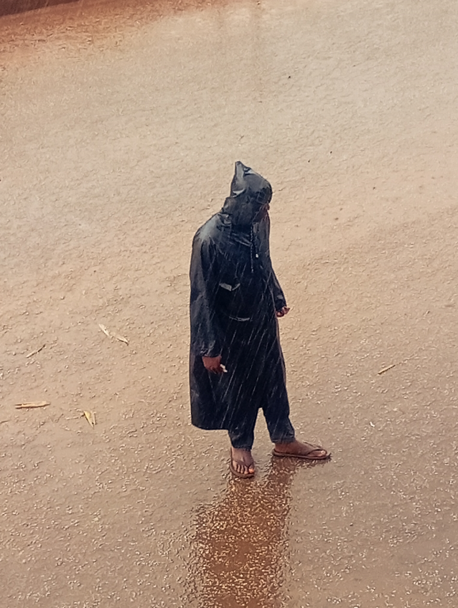 A person wearing a dark, hooded raincoat and flip-flops is standing in the rain on what appears to be a wet, sandy or earthen surface. The person is looking to the right. The scene is outdoors and it is raining heavily. The person is likely located in Bukuru, Nigeria.