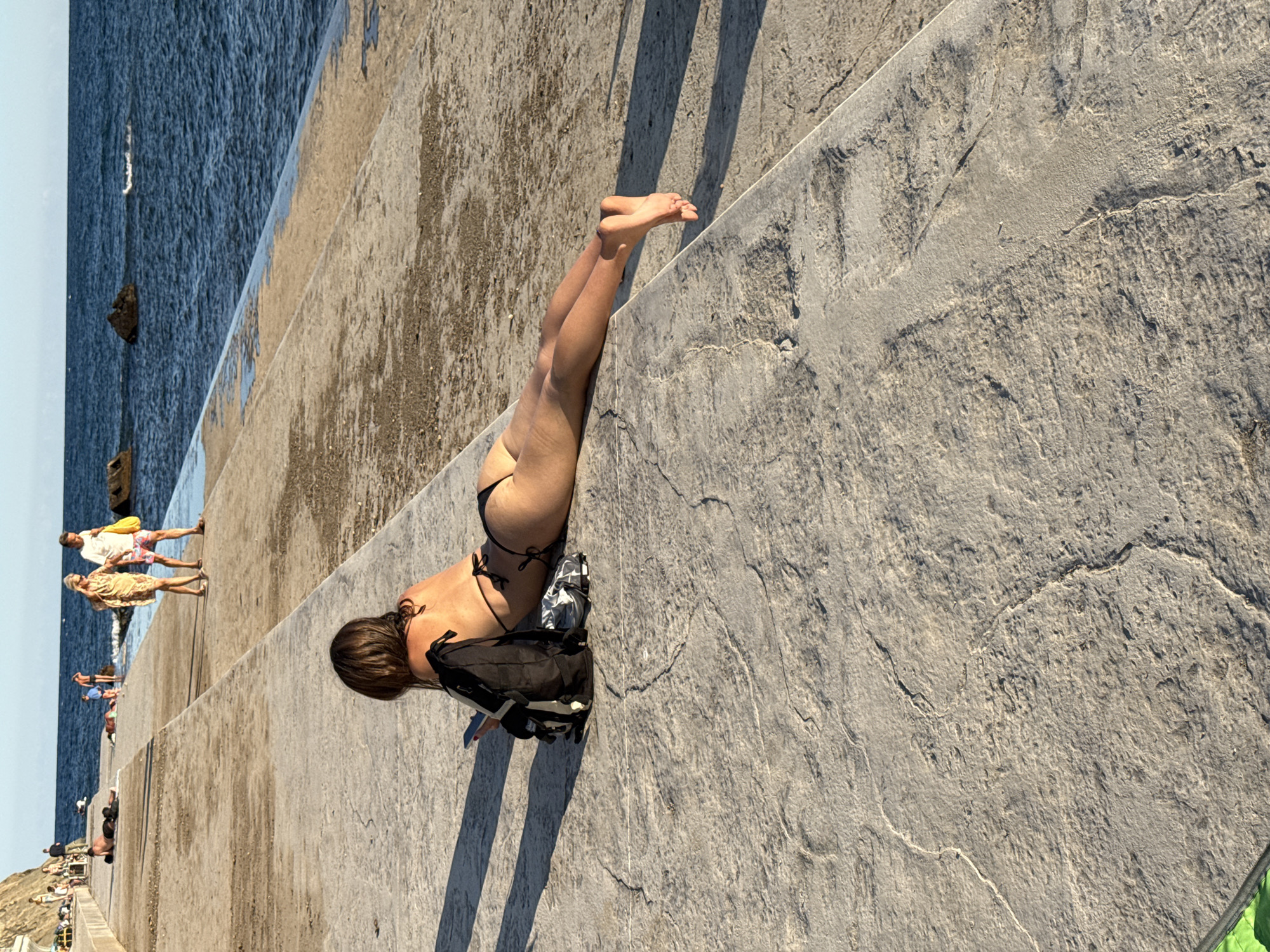 The image depicts a woman in a bikini lying on a concrete walkway next to a body of water. She is facing away from the camera, with her legs extended towards the water. A black backpack and some clothing are placed next to her. In the background, several other people are walking and lounging on the walkway and beach. The ocean is visible in the distance, with a few rocks protruding from the water. The scene appears to be a public area, possibly a promenade or a beach access point. The lighting suggests it is daytime, likely late afternoon or early evening, given the long shadows cast on the walkway. There is no specific information within the image to indicate the location in the city of Ut'os, Ukraine.