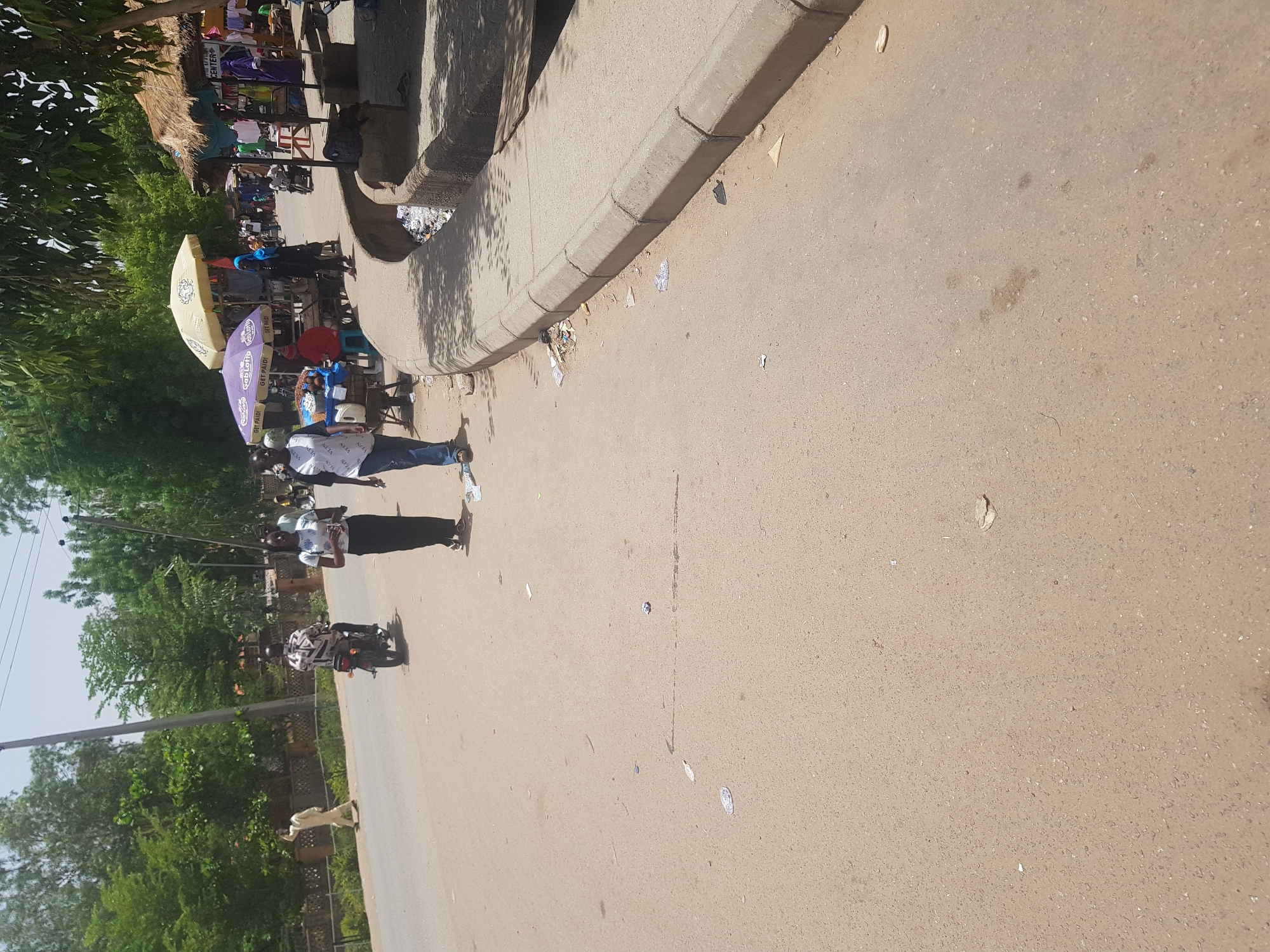 The image depicts a street scene in Dabawa, Nigeria. A concrete curb lines the top-left portion of the frame. A group of pedestrians and vendors are gathered near the curb. One man is standing and wearing a white patterned shirt and dark trousers. A motorcyclist in a black and white patterned shirt is visible further down the road. A light-colored dog is walking in the street, to the left of the motorcycle. Green trees line the left side of the street. The asphalt road is light brown.