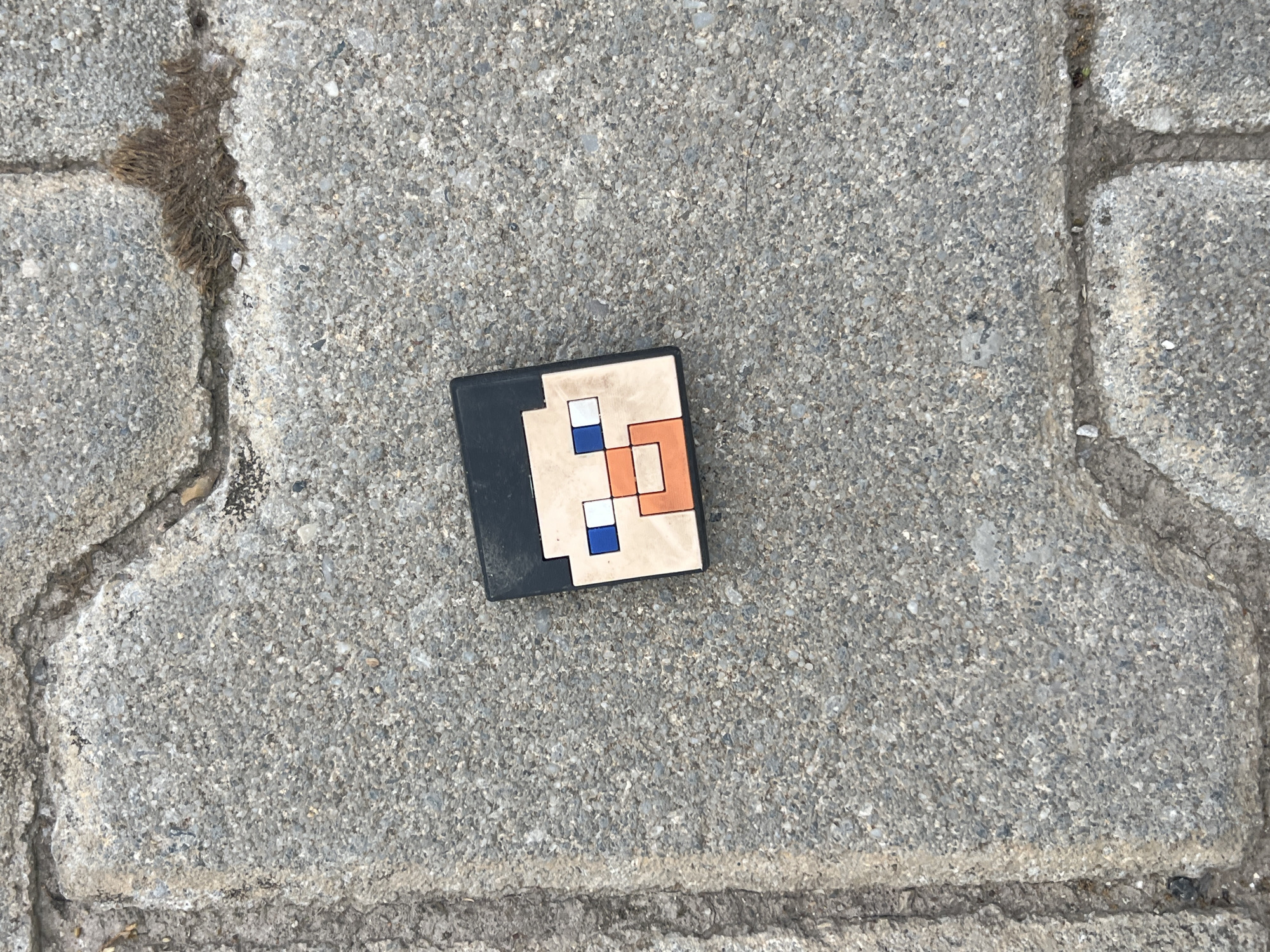 A Minecraft character head toy is lying on a textured concrete surface. The toy has black hair, a beige face with blue eyes, a white pupil in the right eye, and an orange mouth. The concrete appears to be part of a sidewalk or pathway, with visible cracks and aggregate. Some dried organic material is present in the cracks. The image was taken outdoors during daylight. The scene is devoid of any people or discernible urban features, making its specific location within Budva, Montenegro, undeterminable from the visual information provided.