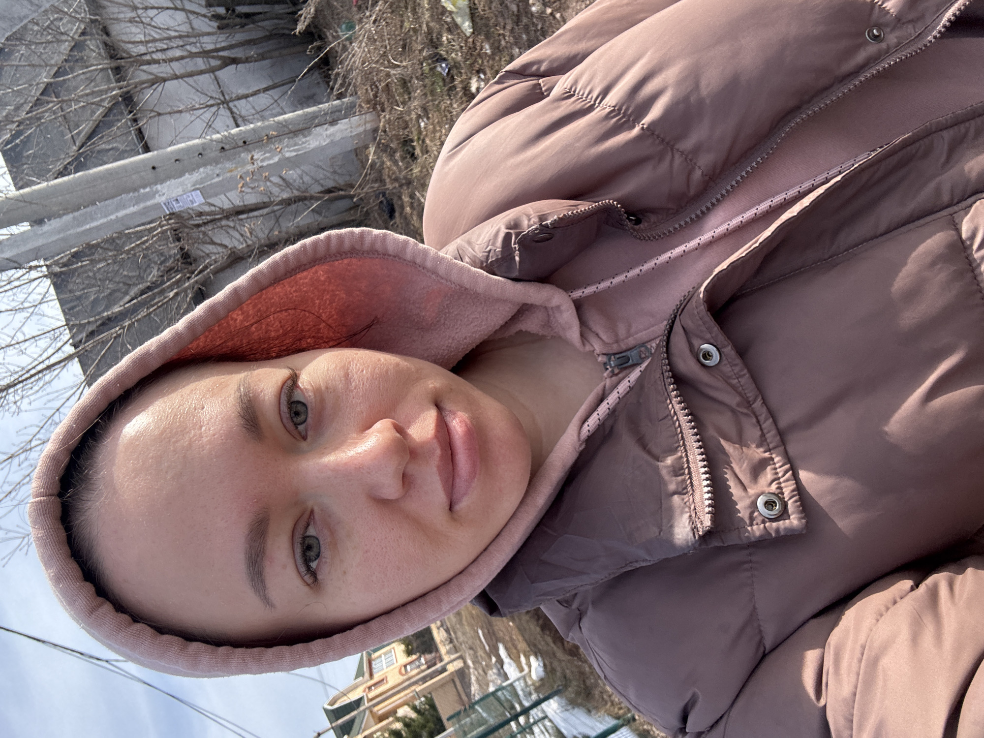 A woman with fair skin, light eyebrows, and green eyes is looking directly at the camera. She wears a light pink hooded sweatshirt under a brown puffer jacket. The hood of the sweatshirt frames her face. The jacket is zipped up partially, exposing the sweatshirt underneath. The background includes a metal guardrail, bare trees, a building with yellow siding and white trim, and a sky. The photo seems to be taken in Ферма, Russia. The lighting suggests it is daytime.