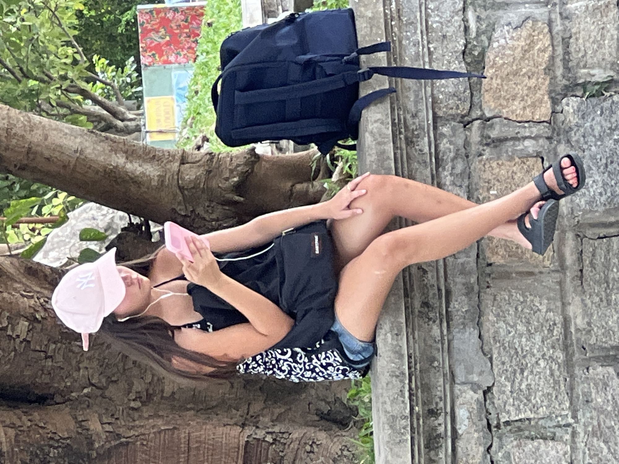 A young woman wearing a pink baseball cap, white earbuds, and a black and white patterned tank top is sitting on a stone wall, looking at her pink smartphone. She is wearing denim shorts and black sandals. A black crossbody bag is draped over her right shoulder, and a large black backpack is placed on the wall next to her. The background consists of a tree trunk, green foliage, and colorful posters or banners. The scene appears to be outdoors, possibly in a park or public area in Đà Nẵng, Vietnam.