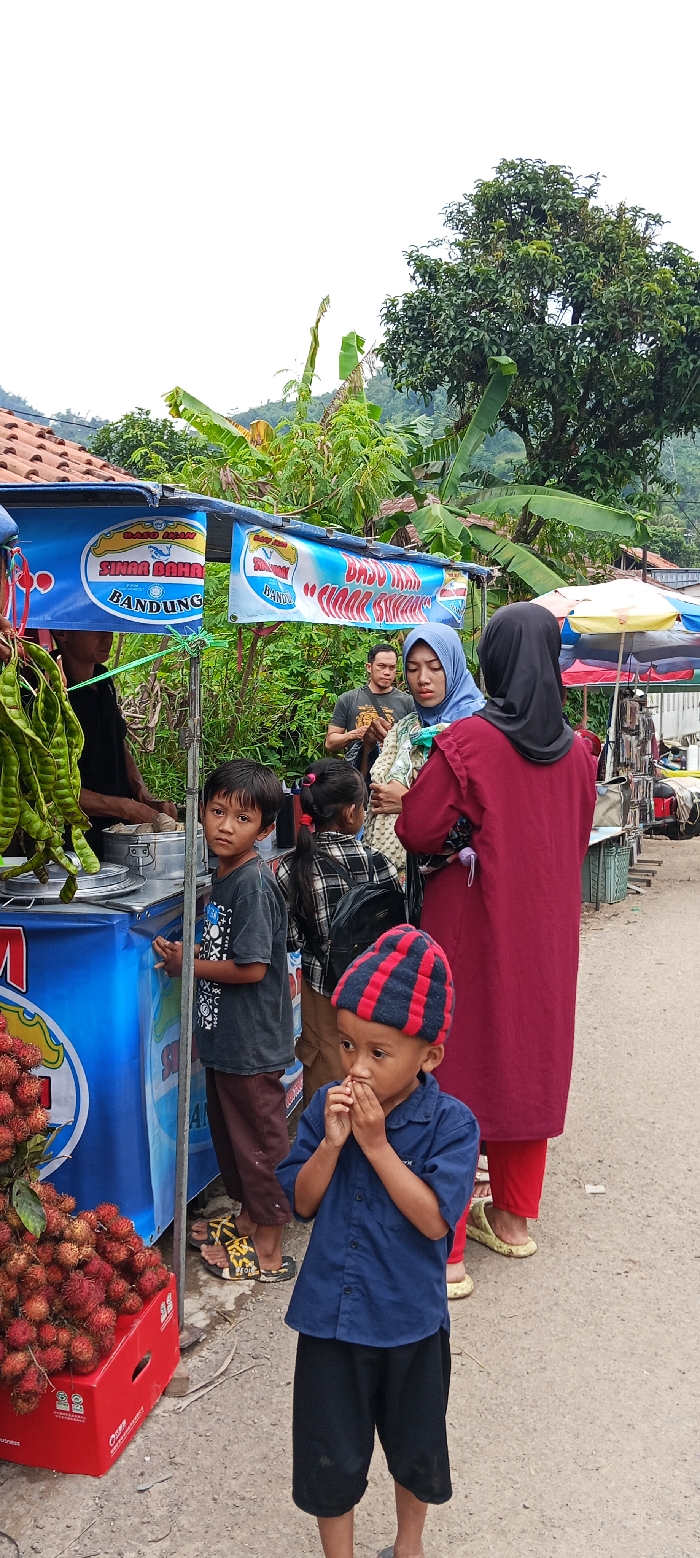 The image shows a street food stall in Kecamatan Kutawaringin, Indonesia. The stall is located at the coordinates -6.973, 107.52, and is identified by two large banners that read “Sina Bahar Bandung” and “Dado Iman Cihaur”. The stall is selling rambutan, a fruit native to Southeast Asia. There are several people at the stall, including a man in a blue shirt, a woman in a red shirt, a woman in a hijab, and three children. The children are all wearing casual clothing. The man is serving food to the woman in the red shirt, while the other two children are watching. One of the children, wearing a black and blue striped hat, is holding his hands together in front of his face. There are a few fruit trees and banana plants visible in the background. The sky is cloudy. The ground is paved with gray asphalt.