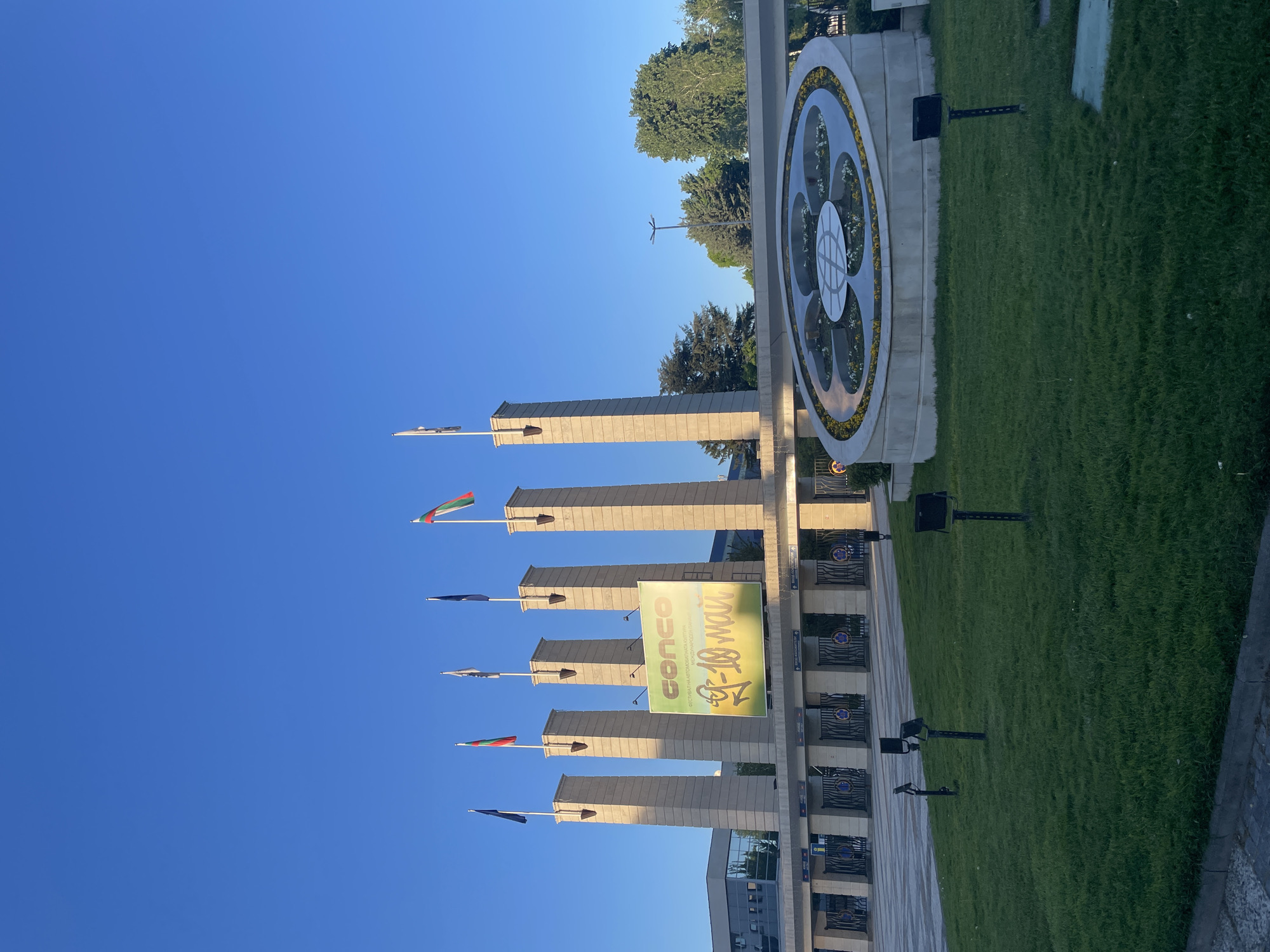 The image shows the Memorial Complex "Defenders of Stara Zagora 1877" in Plovdiv, Bulgaria. The complex is comprised of a series of tall, rectangular concrete pillars, each topped with a flag. A large banner with green text and a date is hung between the pillars. In the background, to the right, there is a circular fountain or monument decorated with flowers. Adjacent to the fountain is a patch of green grass. The sky is clear and blue. The perspective suggests the photo was taken from ground level looking up towards the structures. There are also some smaller black monitors on stands to the right. The ground is tiled.