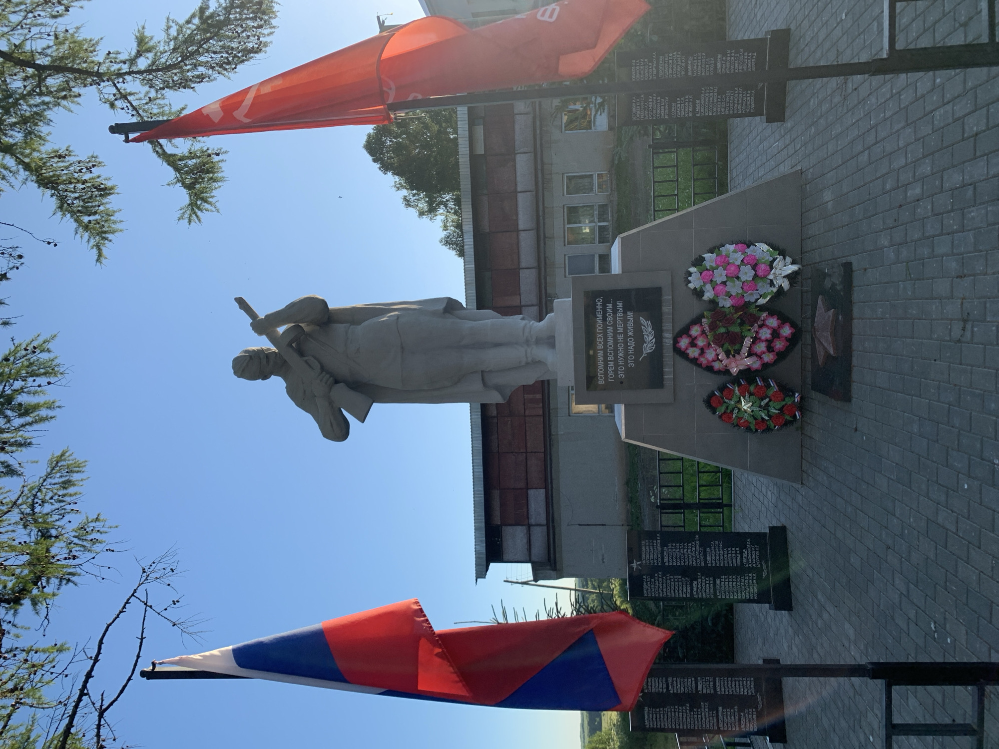 The image shows a war memorial in the city of Svyatitsa, Russia. In the foreground, two flags are displayed: the Russian flag on the left and a red flag, possibly a Soviet or communist flag, on the right. The central element of the memorial is a stone statue of a soldier holding a rifle. Below the statue is a monument with inscribed names and a quote. Wreaths of artificial flowers are placed at the base of the monument. The memorial is set on a paved area with a fence in the background. The environment appears to be outdoors with trees and a clear blue sky. Buildings are visible behind the memorial.