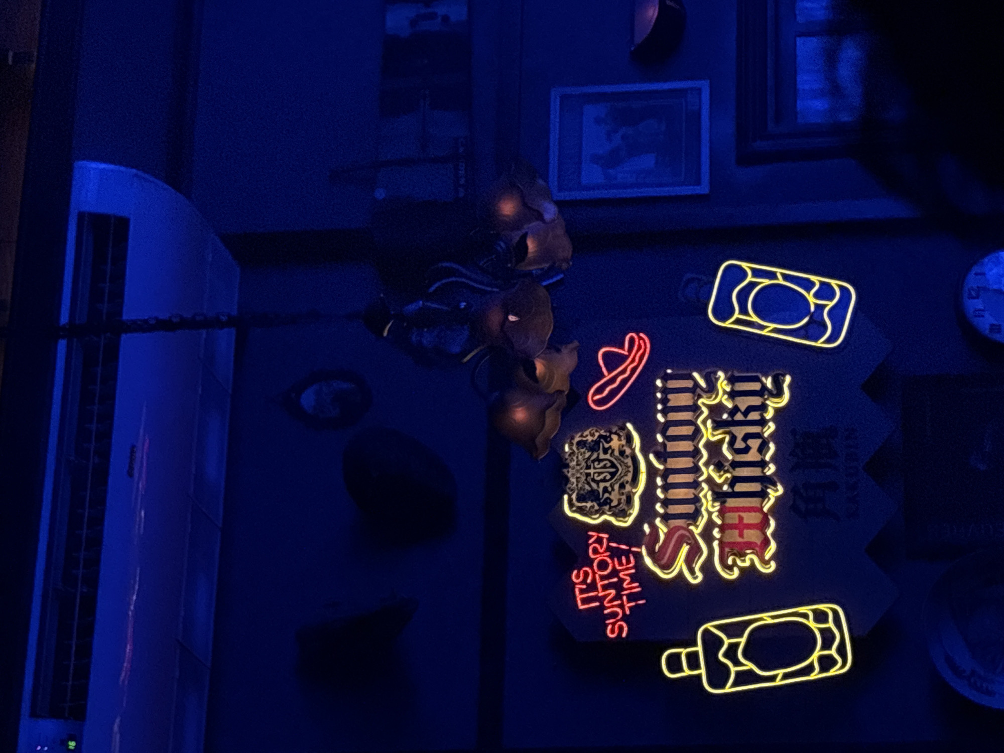 The image depicts a dimly lit interior, possibly a bar or restaurant, bathed in blue and purple light. Dominating the scene is a neon sign that reads "IT'S SUNTORY TIME!" in red lettering, with "Suntory Whisky" written below in yellow. Next to the sign, on the left, is a neon outline of a bottle, and on the right, another neon outline of a bottle with a circular design within. Above the neon sign, a chandelier with multiple illuminated flower-like shades hangs from a chain. Further up, an air conditioning unit is visible on the ceiling. The background features dark walls with framed pictures and some shadowed objects. There are no visible people in the immediate foreground or midground. The overall ambiance is moody and atmospheric, with a focus on the glowing neon signage. The image is taken from a low angle, looking upwards.
