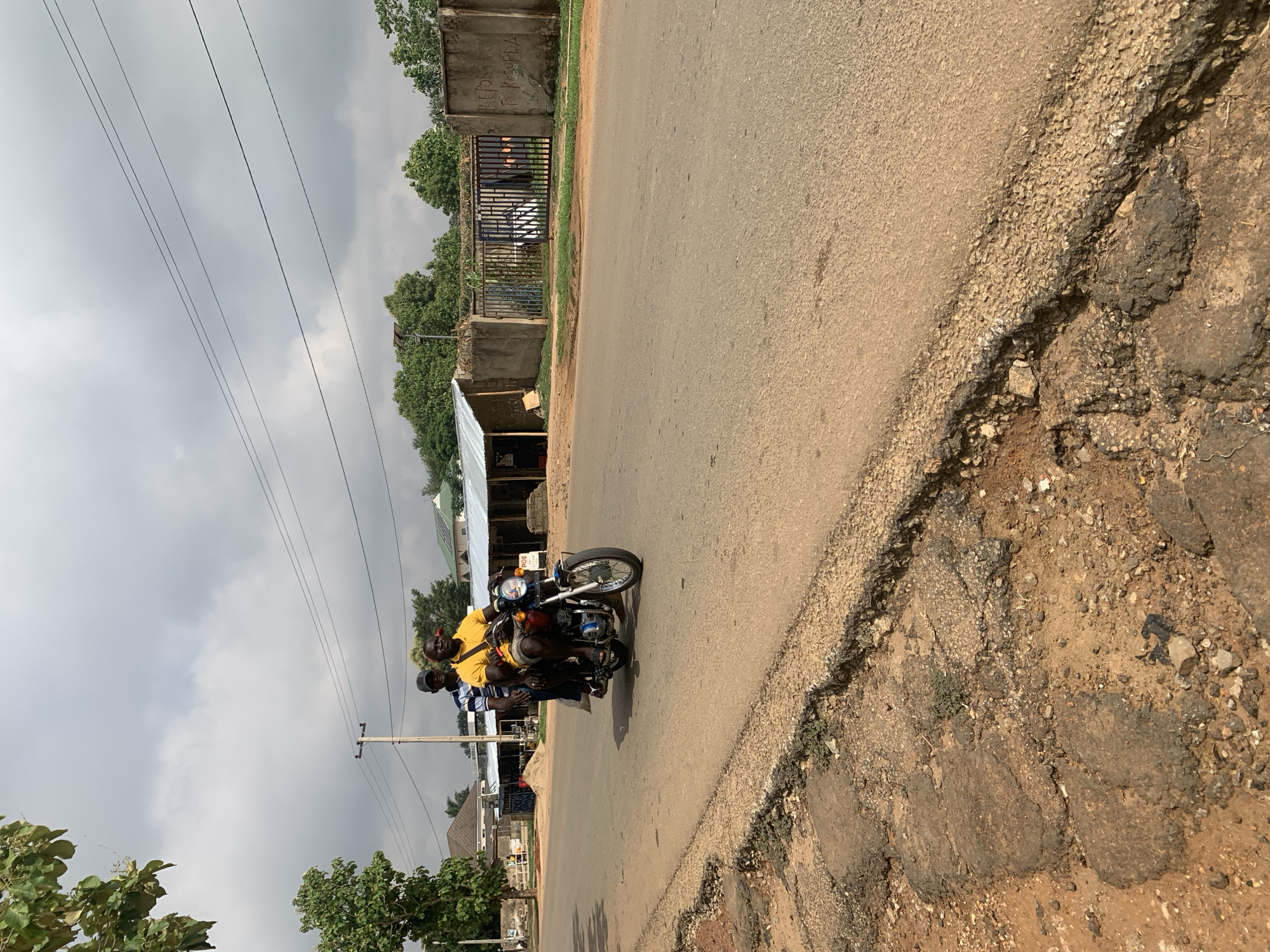 A daytime street scene captured under an overcast sky shows three men riding a motorcycle along a paved road. The driver is wearing a yellow shirt and dark shorts, while the two passengers behind him have striped and darker tops. The motorcycle is black with chrome accents.
The road is in varying conditions; while the main path is paved, its right edge shows significant erosion, transitioning into a dirt and gravel shoulder with visible potholes and cracks. On the left side of the road, there are several simple structures, possibly small shops or residences, with corrugated metal roofs. Further along, a concrete wall and a metal gate are visible. A wooden utility pole stands near the road, supporting multiple power lines that crisscross the gray sky. Green trees and other foliage line both sides of the road and the background, suggesting a semi-urban or rural setting.
On the concrete wall to the left, faded white graffiti is visible, partially reading "REVOLT" and "TRUMP". The overall atmosphere is mundane, depicting daily life and transportation in what appears to be a community in Maigizo, Nigeria.