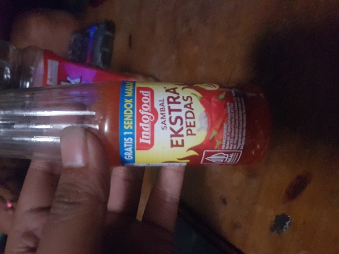 The scene, located indoors in Cimenyan, Indonesia, features a human hand holding a clear glass bottle of Indofood Sambal Ekstra Pedas chili sauce. The bottle contains a red, viscous liquid. Its yellow and blue label displays the brand "Indofood," the product name "SAMBAL EKSTRA PEDAS," the text "GRATIS 1 SENDOK MAKAN," and a Halal certification logo. Illustrations of red chili peppers and garlic are also present on the label. The hand, positioned on the left, shows a single visible fingernail and grips the bottle firmly. The immediate foreground consists of a wooden surface, exhibiting visible grain and minor blemishes. The background is dimly lit and out of focus, revealing a partial view of a dark pink or red container and an unidentified dark rectangular object.