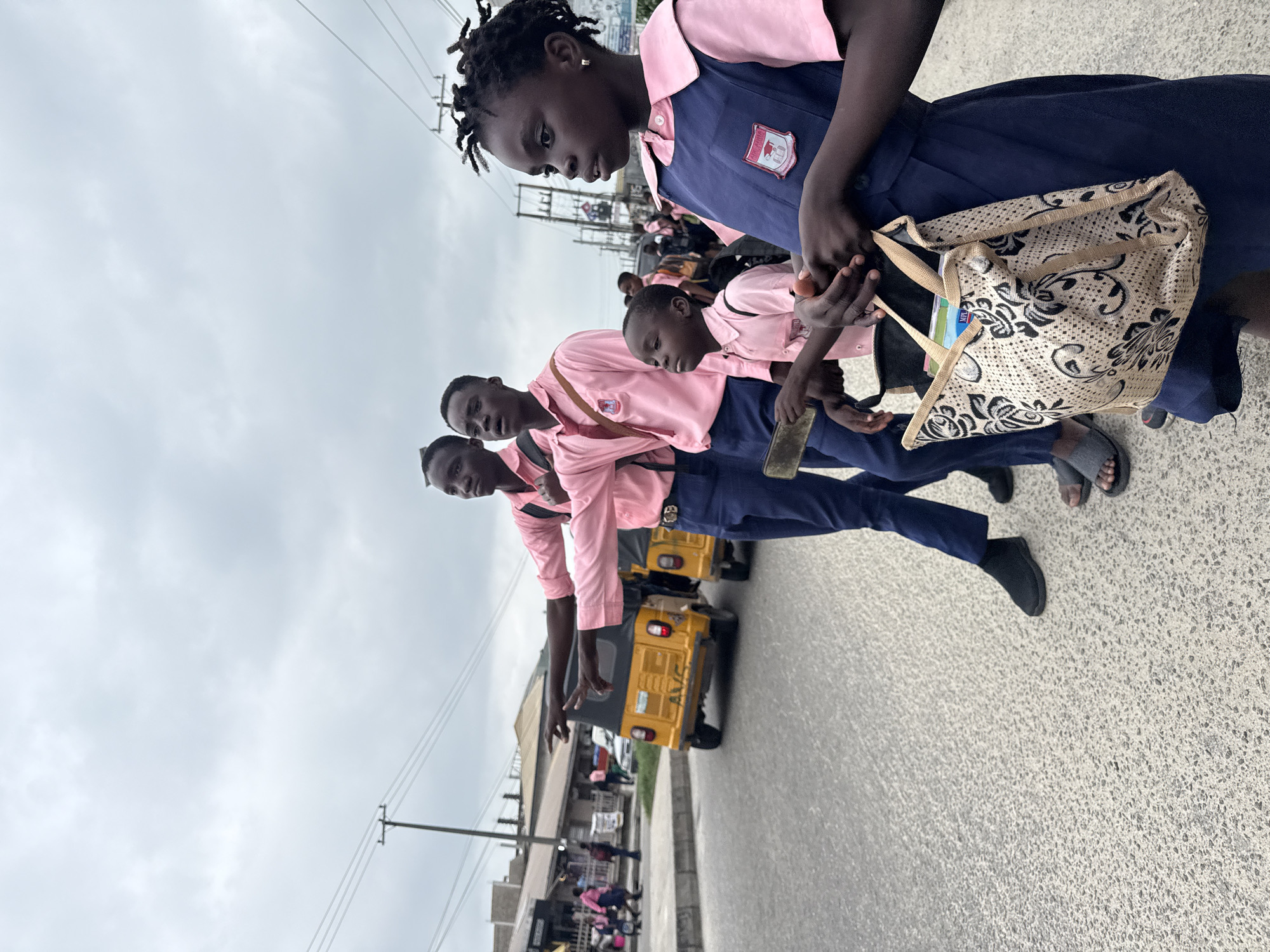In Ado, Nigeria, on a cloudy day, a group of schoolchildren in uniform stand on a paved street. The foreground is dominated by a young girl wearing a navy blue pinafore over a pink collared shirt, carrying a patterned tote bag. Behind her, three boys in similar pink shirts and navy trousers stand. The two older boys on the left are pointing to the left, with one holding a flat object. In the background, a yellow auto-rickshaw, a common mode of transport in Nigerian cities, is visible on the street. Beyond that, buildings and a telephone pole line the street, suggesting an urban environment. The sky is overcast, casting a diffused light on the scene.