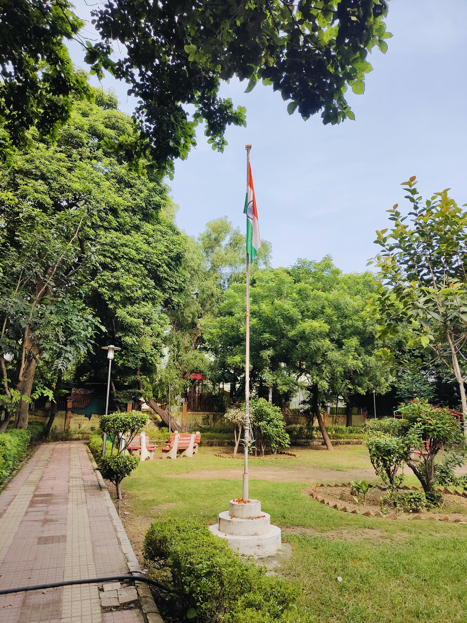 An outdoor scene in Gurgaon, India, features a park with lush greenery and a paved walkway. In the foreground, a red and beige tiled walkway runs from the left side of the frame towards the background. To the right of the walkway, a well-maintained lawn is dotted with manicured bushes. A flagpole stands prominently in the center of the lawn, flying the Indian flag. Behind the flagpole, a white concrete base with a tiered structure is visible, adorned with small orange flowers. Further back, several red benches are placed on the grass, and mature trees with dense foliage provide shade. The background reveals more trees and a clear sky. The overall atmosphere is peaceful and natural.