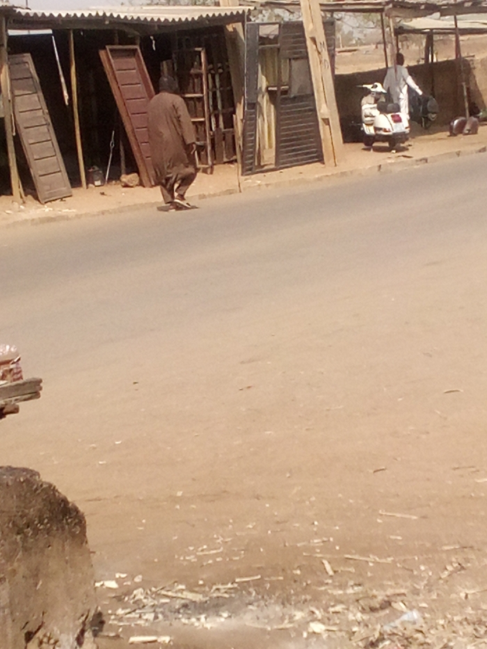 This image depicts a daytime street scene in Tudun Murtala, Nigeria. The foreground shows a dusty, light brown dirt shoulder with scattered white debris. A paved, grey-brown road runs horizontally across the midground.
In the background, on the left, a man wearing a long, dark brown traditional garment and dark headwear is walking away from the camera, moving towards the right side of the frame. He wears dark open-toed sandals. Behind him is an open-fronted workshop or stall with a corrugated metal roof supported by wooden poles. Inside and around this structure, multiple brown wooden door panels or shutters are visible, some leaning against walls, indicating a carpentry or door-making business.
Further to the right in the background, another man in a light-colored long-sleeved top and trousers is standing near a white scooter, possibly a Vespa, which is parked on the dirt shoulder. The man appears to be facing or attending to the scooter or an object behind it. Behind him, additional rudimentary structures with roofs are visible. Two light-brown animals, possibly goats, are on the ground near these structures on the far right. The overall environment suggests a roadside commercial area.